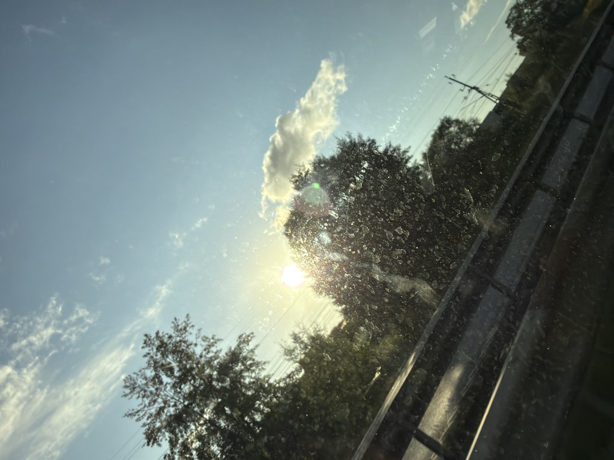 The image depicts a view through a dirty window, likely from a vehicle, looking towards a bright, sunlit sky. The sun is intensely bright, positioned slightly to the left of the center, casting a strong glare and lens flare. Silhouetted trees with dense foliage are visible against the sky, some of which are illuminated by the sunlight. Wispy white clouds are scattered across the clear blue sky. A power line pole with visible wires is present in the background, suggesting an urban or suburban environment. A dark railing or barrier is prominent in the foreground, running horizontally across the lower portion of the image. The overall scene suggests a daytime setting, possibly late afternoon due to the low angle of the sun and the warm lighting. The presence of the power pole and what appears to be a road barrier indicates a location along a transportation route. The dirt on the window obscures some details of the scene. There are no people or specific identifiable landmarks that would pinpoint a precise location within Mölndal, Sweden.