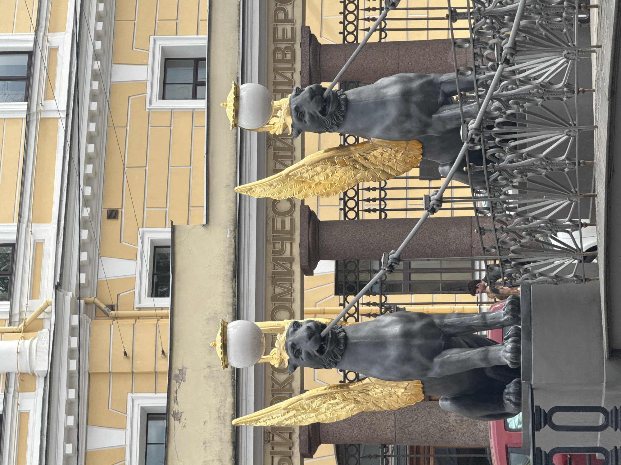 The image depicts two griffin statues, likely part of the University Embankment railings in Saint Petersburg, Russia. The griffins are dark gray with golden wings, each holding a spear-like pole topped with a spherical lamp. They are positioned in front of a building with a yellow facade and white window frames. The building's inscription includes the word "УНИВЕРСИТЕТ". A decorative metal fence is also visible. Several people can be seen indistinctly in the background. The shot is taken at eye level. A red car is visible partially on the lower right.
