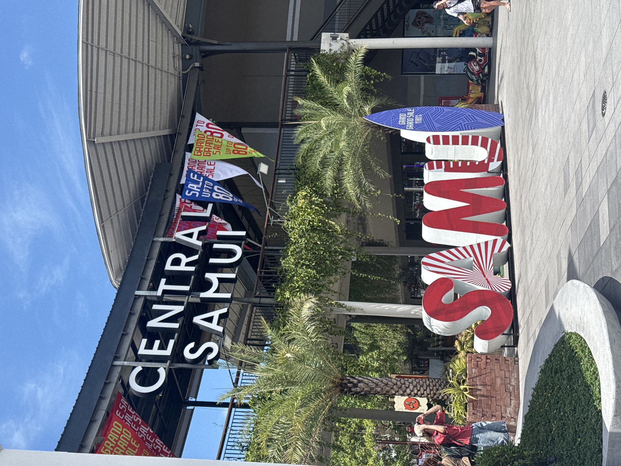 In Ko Samui District, Thailand, a large outdoor sign spelling "CENTRAL SAMUI" is prominently displayed. The sign is mounted above the entrance to a building with a modern, sloping roof. Various banners advertising a "GRAND SALE UPTO 80%" hang from the building's facade. In front of the building, a massive 3D sculpture spelling "SAMUI" is visible, with the letters "S" and "A" decorated in red and white stripes, and the letter "U" replaced by a blue surfboard with the "GRAND GRAND SALE 80%" text. Palm trees and lush green hanging plants surround the area, adding to the tropical ambiance. Several people are present: one individual in the foreground is taking a photo with their phone, another person is walking in the background. The environment appears to be a pedestrian plaza or walkway, with patterned paving stones and a curved concrete planter filled with greenery. The sky is clear and blue, suggesting a sunny day.