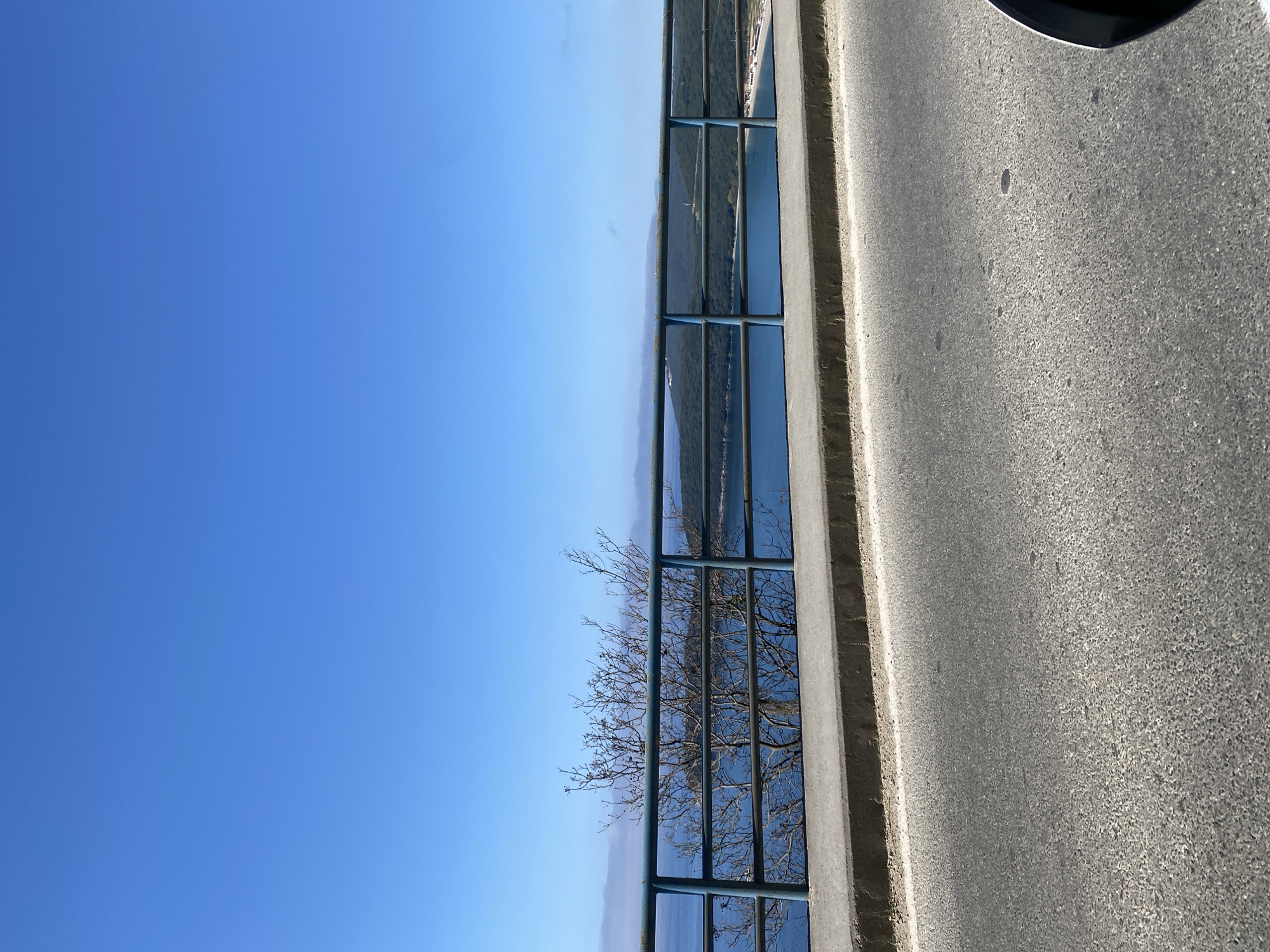 The image shows a view from a road in Prijevor, Montenegro. The right side of the image features asphalt road. A concrete barrier with a metal fence runs along the edge of the road. Beyond the fence is a view of water and distant land, partly obscured by a bare tree branch. The sky is clear and blue.