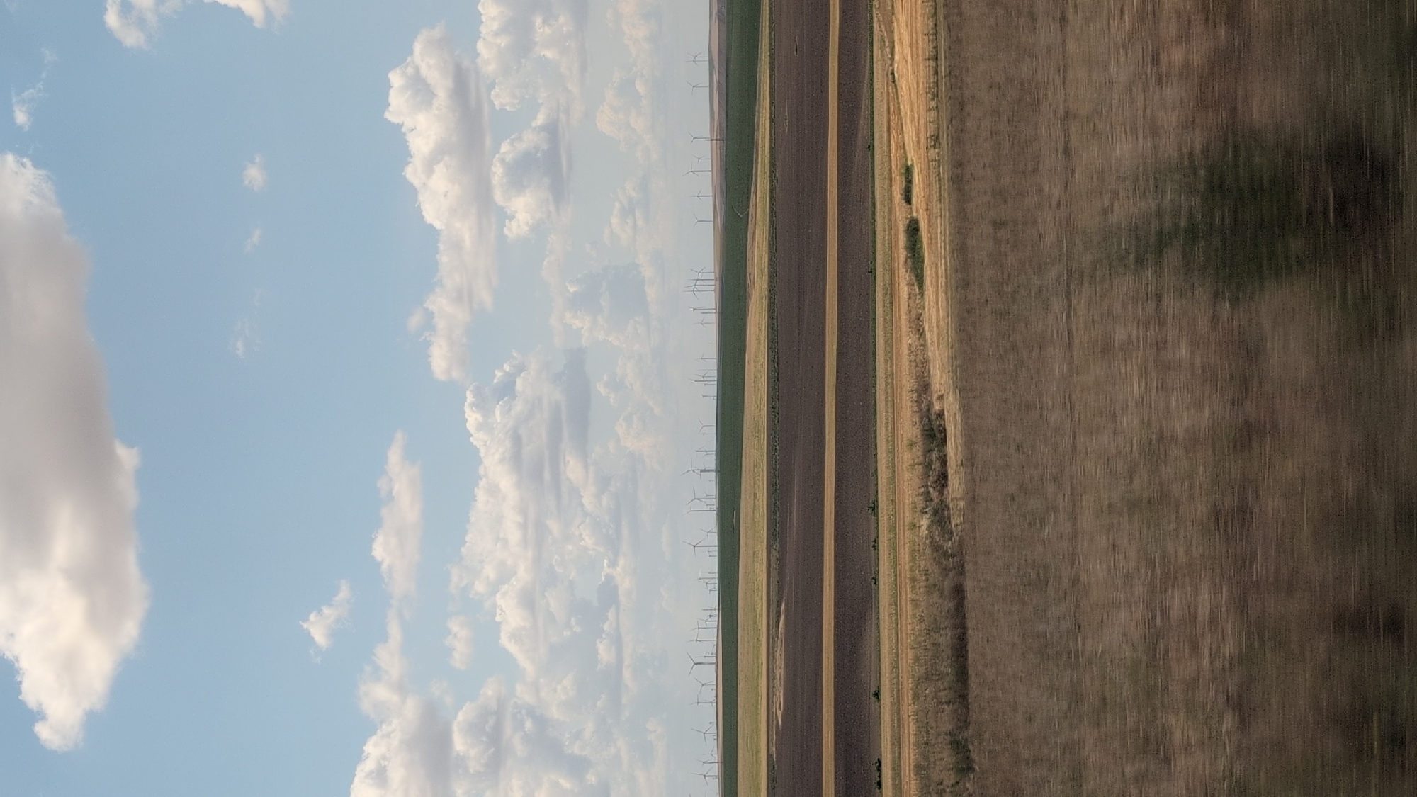 The image depicts a vast agricultural landscape under a partly cloudy sky. In the foreground, the fields are blurred due to motion, suggesting the photo was taken from a moving vehicle. The middle ground shows distinct rectangular plots of land, some appearing plowed and dark brown, others with dry, yellowish vegetation. In the distance, a large wind farm stretches across a green, rolling hill, with numerous wind turbines clearly visible. The sky is a soft blue with scattered white and gray clouds. The scene is characteristic of rural areas with renewable energy infrastructure. There is no discernible indication of the city of Baia, Romania, within the image.