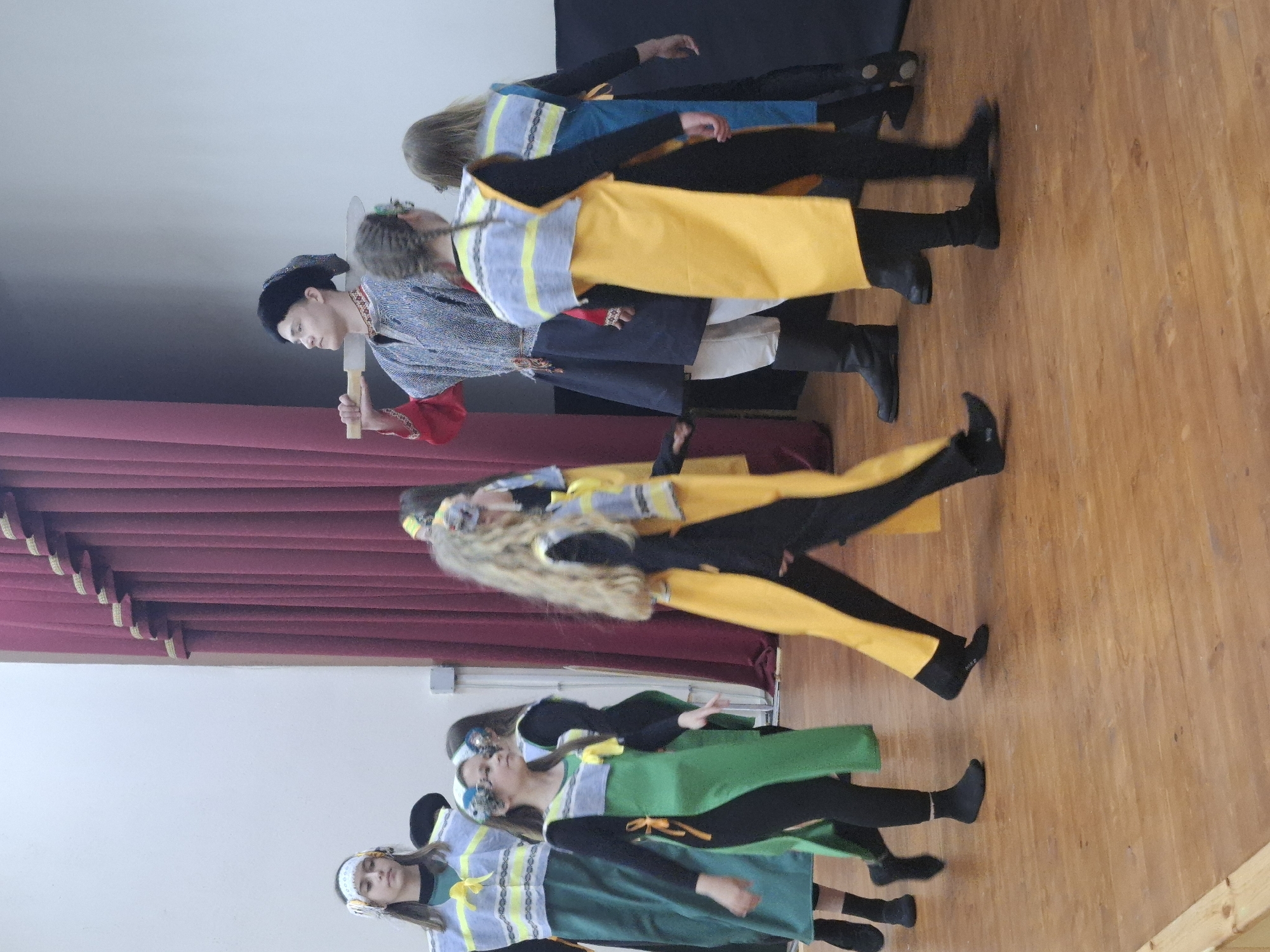 A staged performance is captured in an indoor setting, likely a theater or auditorium, in the city of Марковский, Russia. A line of individuals are dressed in costumes of blue, yellow, and green. A male figure stands on stage in traditional attire, holding a prop, possibly an axe. The backdrop consists of a dark curtain, a wall, and a wooden floor.