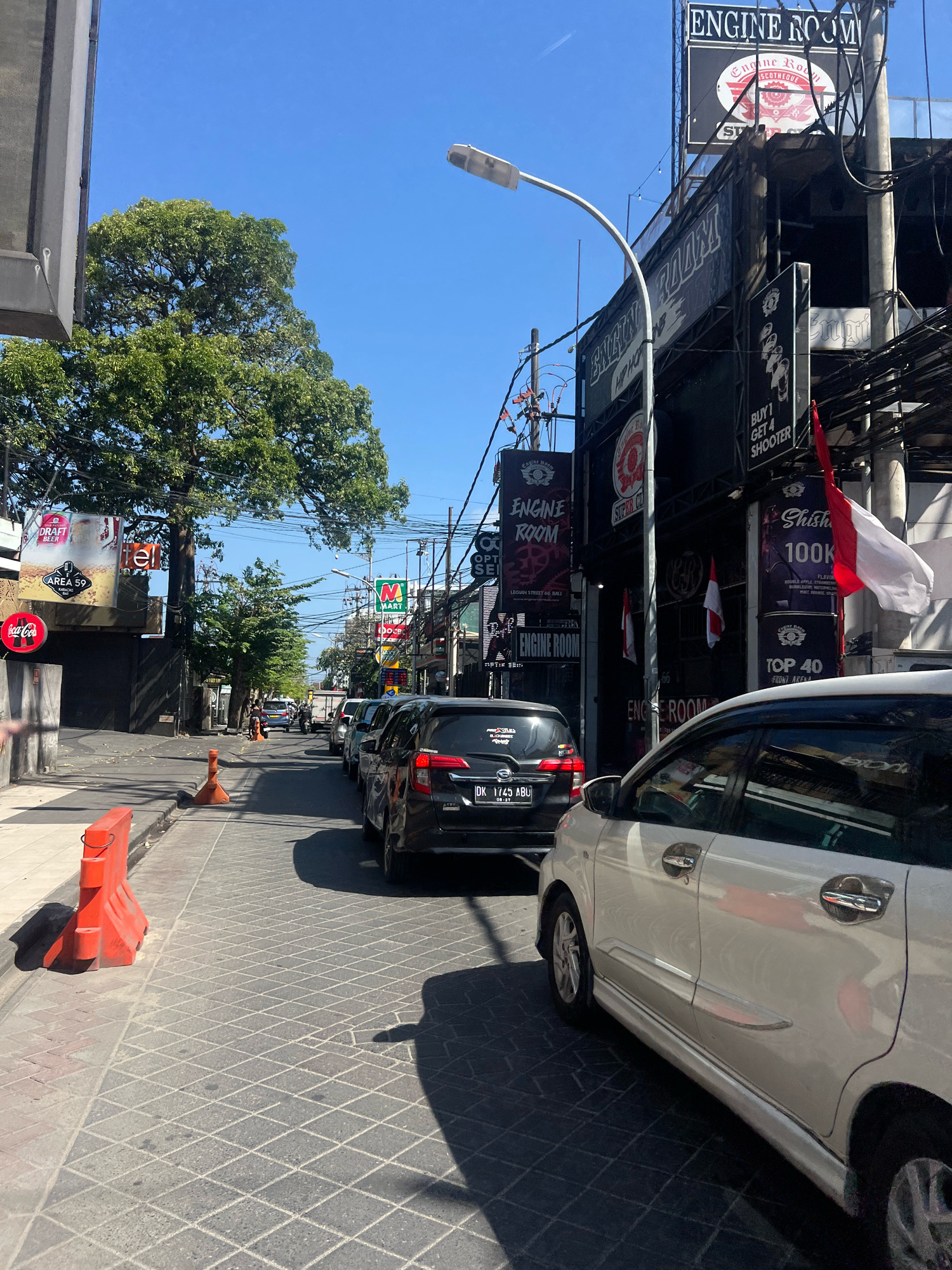 The image depicts a street scene in Kuta, Indonesia, under a clear blue sky. The street is paved with interlocking stones and is lined with buildings and trees. A traffic jam of cars is visible, with several vehicles stopped or moving slowly. On the left side of the street, there are orange traffic barriers and a large tree. Buildings on the right side of the street feature multiple signs advertising "ENGINE ROOM," "DRAFT BEER," and "M Mart." One sign indicates "BUY 1 GET 4 SHOOTER," and another displays "Shisha 100K." A white car is parked in the foreground on the right, and a black SUV is in front of it, partially obscuring it. Indonesian flags are displayed on poles attached to the buildings. The overall impression is of a busy commercial street in a tropical urban environment.