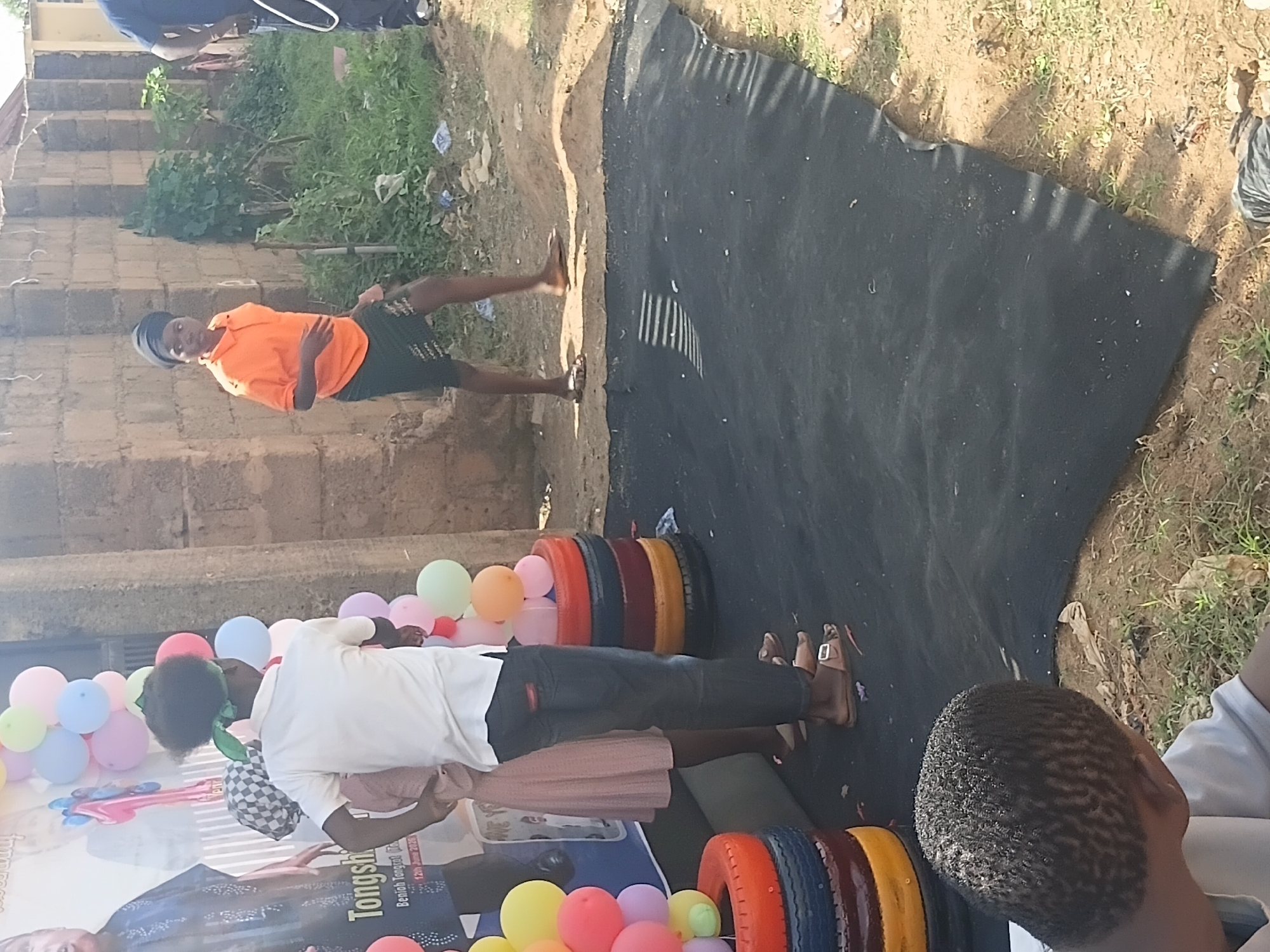 The image depicts an outdoor scene, likely in the city of Jos, Nigeria, possibly during a celebration or event. In the foreground, a person is seen from the back, wearing a white t-shirt and dark pants, with their back to the camera. To their left, another person is partially visible, wearing a light pink dress.
In the midground, a young person wearing an orange t-shirt and a dark patterned skirt stands facing left. Behind them, a brick wall forms a backdrop, with some vegetation growing nearby. Colorful balloons are arranged in clusters, some floating above stacks of painted tires which appear to be used as decoration or seating.
In the background, the brick wall continues, with hints of buildings and a fence. The ground is a mixture of dirt and a black fabric covering, possibly a mat or a tarp. The overall impression is of a lively outdoor gathering.
