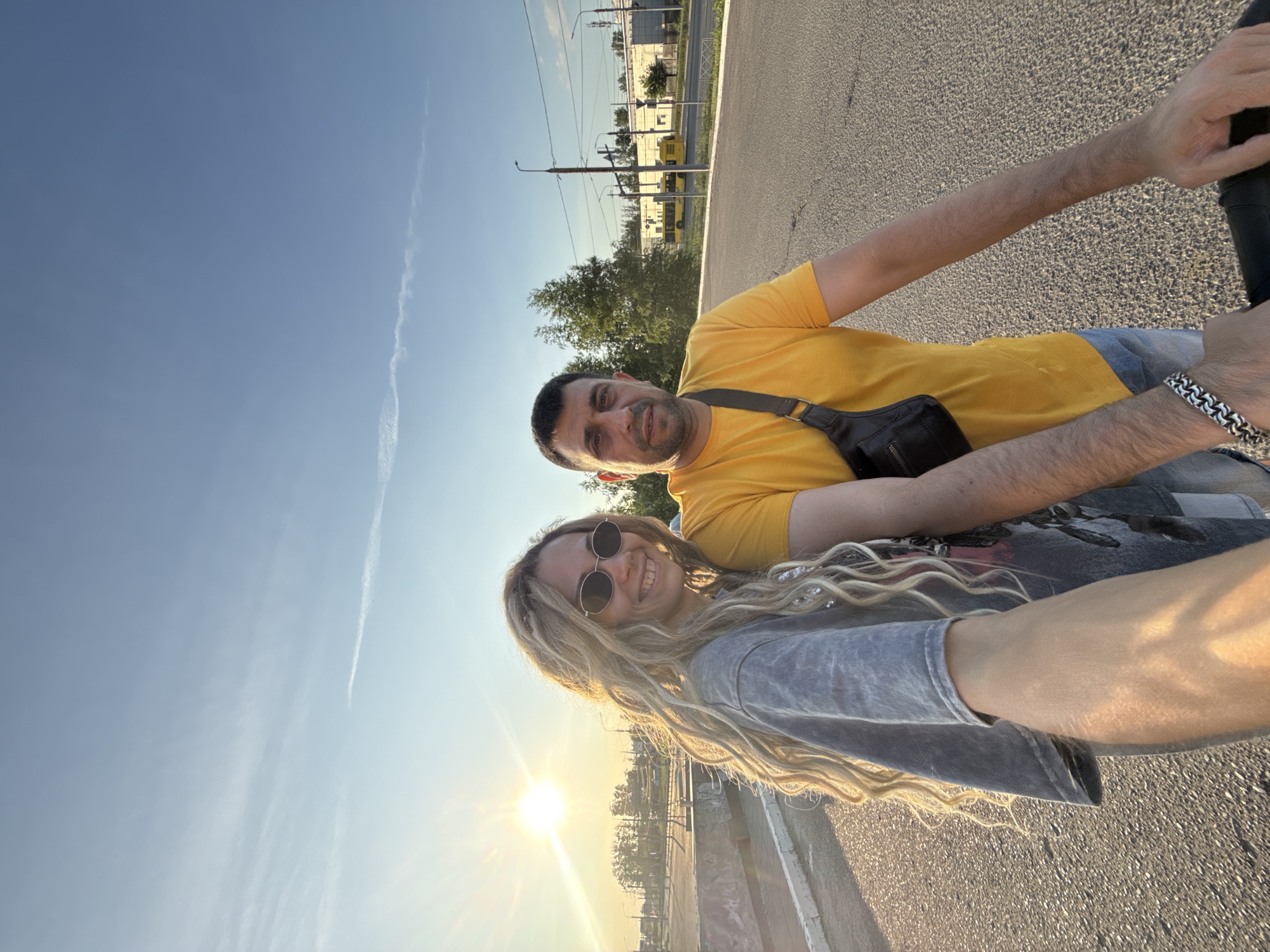 A full-shot, outdoor, eye-level, selfie-style photograph captures two individuals, a woman and a man, standing side-by-side on an asphalt road with a clear blue sky and some wispy clouds overhead. The sun is low in the sky, creating a warm, golden-hour glow that illuminates the scene and casts long shadows.
The woman, on the left, has long, blonde, braided hair and is wearing sunglasses and a grey t-shirt with a faded graphic print. She is smiling and her left arm is extended, holding the camera.
The man, on the right, has short, dark hair and is wearing a bright yellow t-shirt and blue jeans. He has a black fanny pack slung across his chest. He is looking directly at the camera with a neutral expression.
In the background, the environment appears to be an urban or semi-urban setting. There is a sidewalk or paved area to the left of the road, with some low-rise structures and greenery beyond. In the middle ground, there are trees and buildings, including a yellow bus visible on what looks like a street or highway. Utility poles and wires are also present. The overall impression is of a casual, daytime outing in a Russian city, possibly Заволжье, Russia, given the architecture and the presence of a public transport bus that appears to be characteristic of the region. The road surface is asphalt, and there are some white lines marking the edge of the roadway.