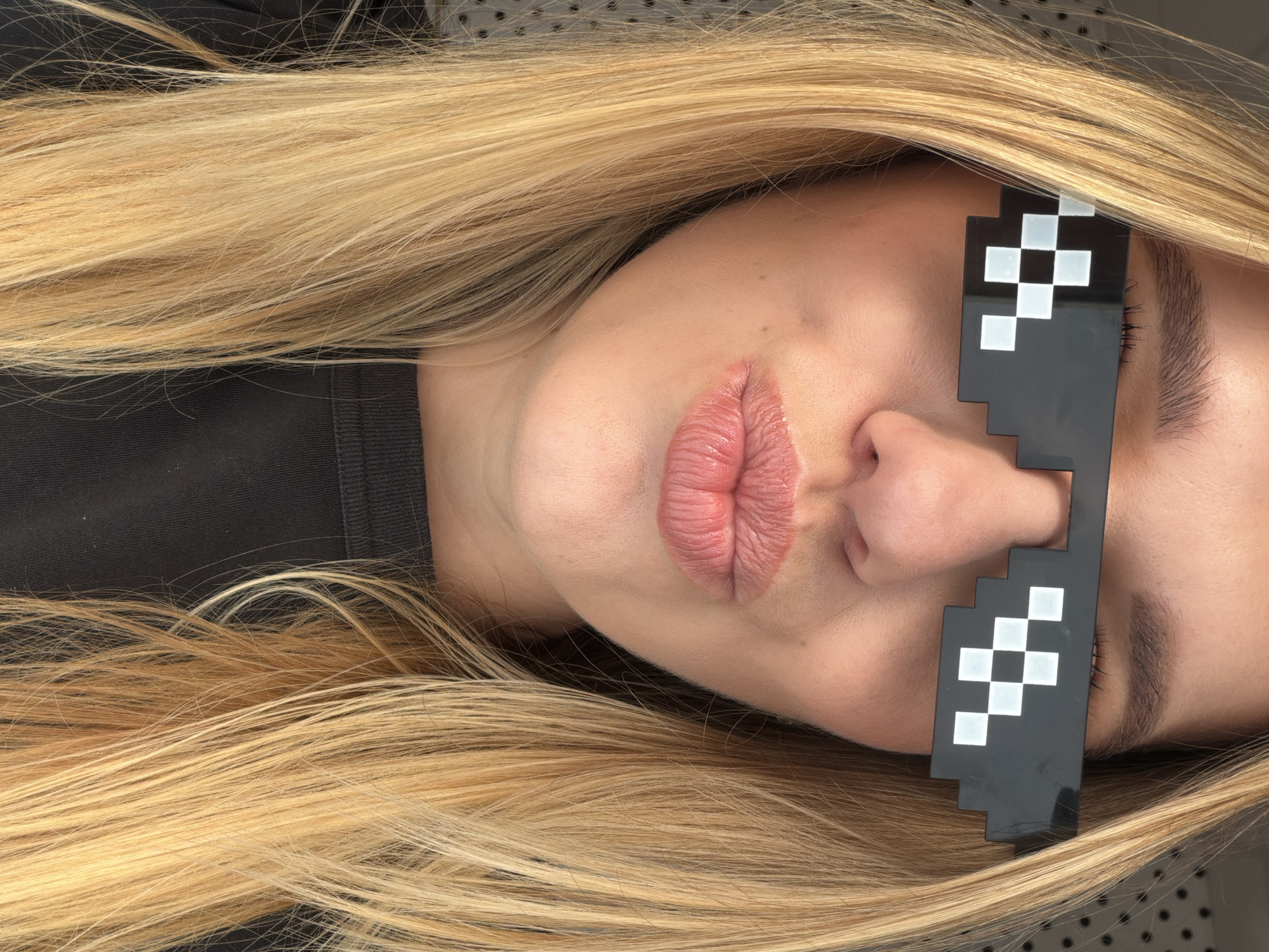 A fair-skinned person lies with their head to the right, facing upwards. They have long, blonde hair spread around their head and shoulders. The person is wearing pixelated, black sunglasses with white squares. The background appears to be a textured grey surface. The clothing worn is a dark color. The image provides insufficient information to definitively locate the scene in Riga, Latvia, or to confirm that it is even there.