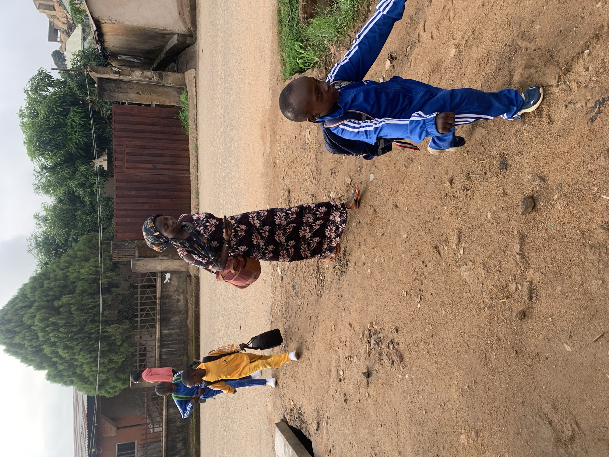 This media file captures a group of people walking along a street in Jos, Nigeria. The scene is characterized by a dirt path on the right that transitions into a paved road on the left, with residential buildings and lush greenery forming the background.
In the foreground, an adult woman, dressed in a long, dark floral-patterned dress and a headscarf, walks on the dirt path. She carries a woven handbag in her left hand and wears sandals. Adjacent to her and slightly ahead, a young boy in a bright blue tracksuit with white stripes on the sleeves and legs carries a dark backpack. He is wearing sneakers.
Further along the paved section of the street, another boy, clad in a yellow tracksuit and carrying a backpack, is visible. Behind him, two more children are partially seen: one in a blue top and another in a pink top. The children's attire and backpacks suggest they may be on their way to or from school.
The background features various buildings, some with corrugated metal roofs, indicating a residential area. A dense green tree stands prominently, along with a brown corrugated metal fence and concrete wall structures. The sky is overcast, suggesting cloudy weather during the daytime. No visible text is discernible in the image. The overall setting depicts a typical street scene in a semi-urban or residential area of Nigeria.
