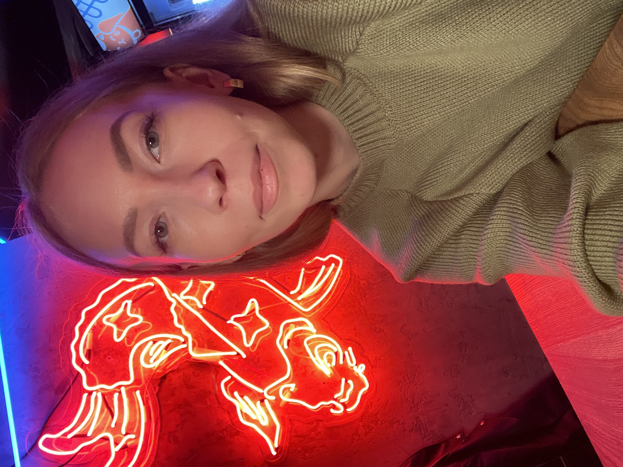 A woman with blonde hair is posing for a selfie in front of a neon sign. The sign appears to be red and depicts a stylized creature, possibly a sea creature or mythical being with stars, against a textured wall. To the left of the red neon sign, there is a blue neon light. The woman is wearing a knitted green sweater. The location is in Perm, Russia.