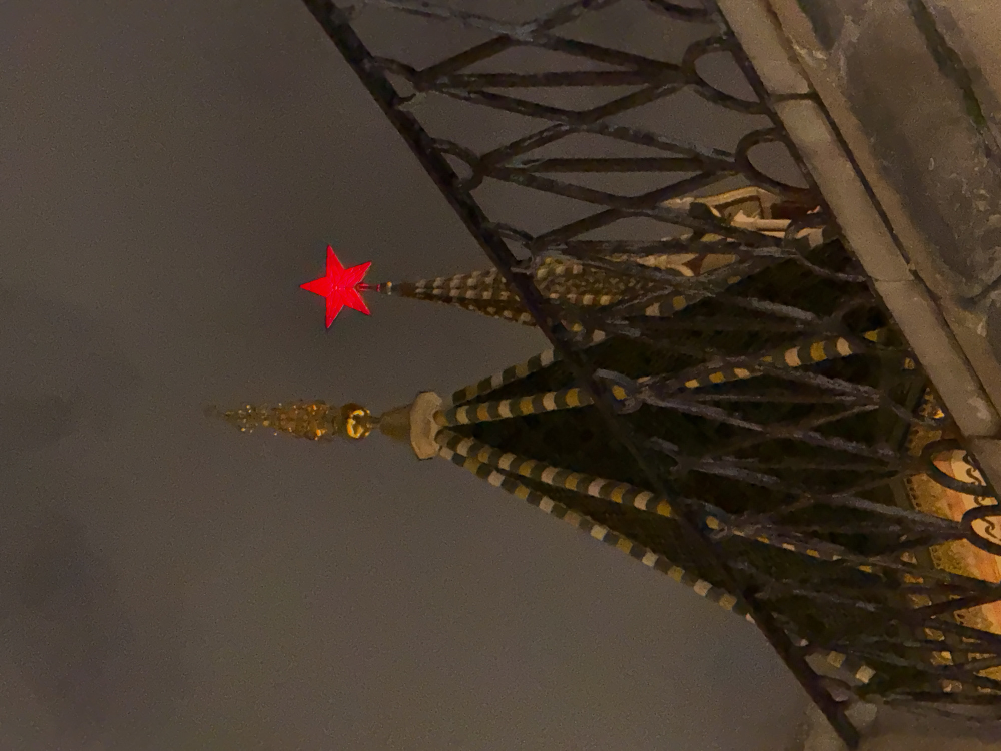 The image shows a nighttime view of one of the towers of St. Basil's Cathedral in Moscow, Russia. The tower is topped with a red star, which is illuminated. A decorative metal railing is visible in the foreground. The sky is dark and overcast. The scene is viewed from below, looking upwards.