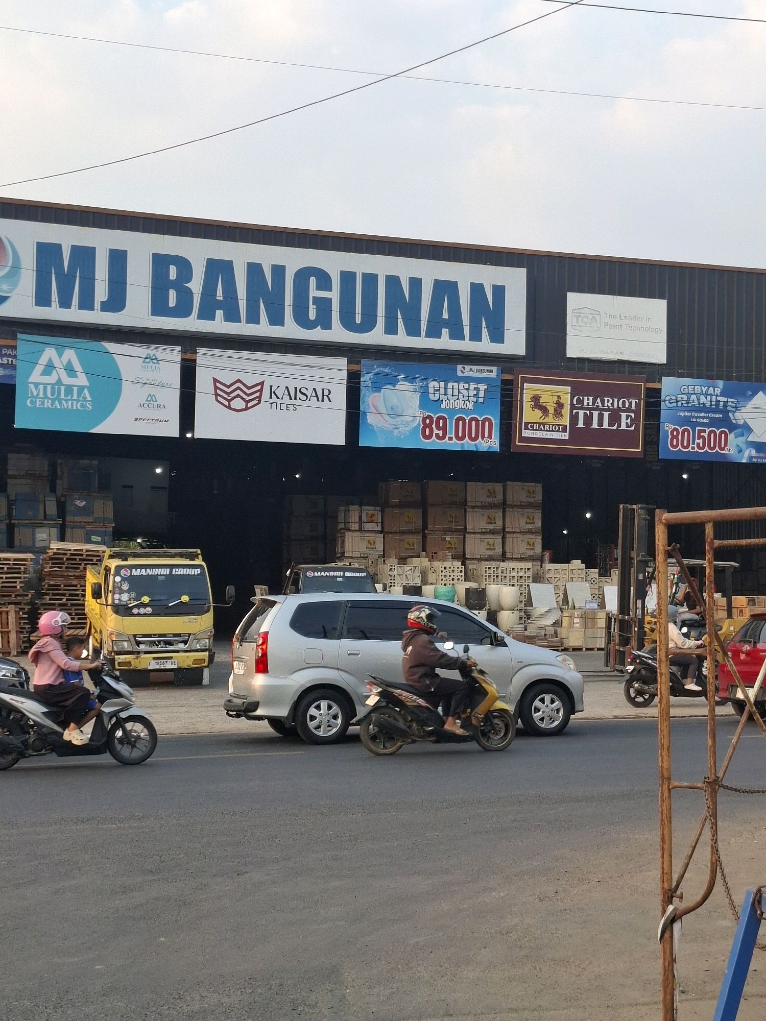 The image captures a street view in front of a building identified as "MJ BANGUNAN" in Karangtengah, Indonesia. The building appears to be a large construction supply store, as indicated by the various advertisements for tiles and building materials displayed on its facade. These advertisements include "MULIA CERAMICS," "KAISAR TILES," "CHART TILE," and "GEBYAR GRANITE."
In the foreground, a paved road is visible with several vehicles. A silver minivan is positioned centrally, and to its right, a red car is partially visible. Multiple motorcycles are also present. One motorcycle, ridden by a person wearing a pink headscarf and a dark-colored jacket, is moving towards the left of the frame, with a passenger behind them. Another motorcycle, ridden by a person wearing a brown jacket and a helmet, is moving towards the right of the frame.
Behind the vehicles, the entrance to the "MJ BANGUNAN" store is visible, with stacks of building materials, including what appear to be pallets of tiles and concrete blocks, visible through the open doorway. A yellow truck with "MANDIRI GROUP" written on its side is parked to the left of the entrance. The sky above is partly cloudy. A metal structure, possibly part of a temporary fence or scaffolding, is present on the right side of the frame.