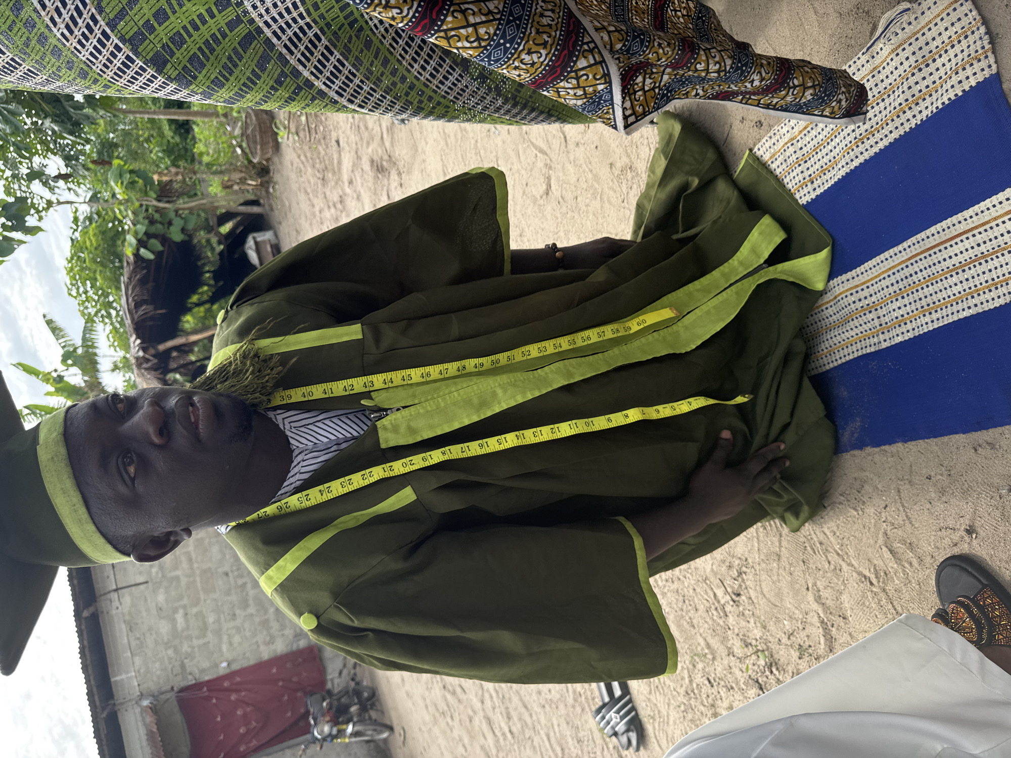 A man wearing a green robe with yellow trim is being measured with a yellow tape measure around his neck and chest, presumably for tailoring. He also wears a matching green cap. The photo appears to be taken outdoors in Tofa, Nigeria, judging by the sandy ground, simple structures visible in the background, and the attire of the people around him. There are two other individuals visible only from the torso up, one wearing a fabric with a green and black geometric pattern and the other wearing a colorful patterned fabric. A pair of sandals lies next to the green robe on the sand. A striped mat lies near the robe as well. In the background, a motorcycle is parked near a weathered building, adding to the candid nature of the scene.