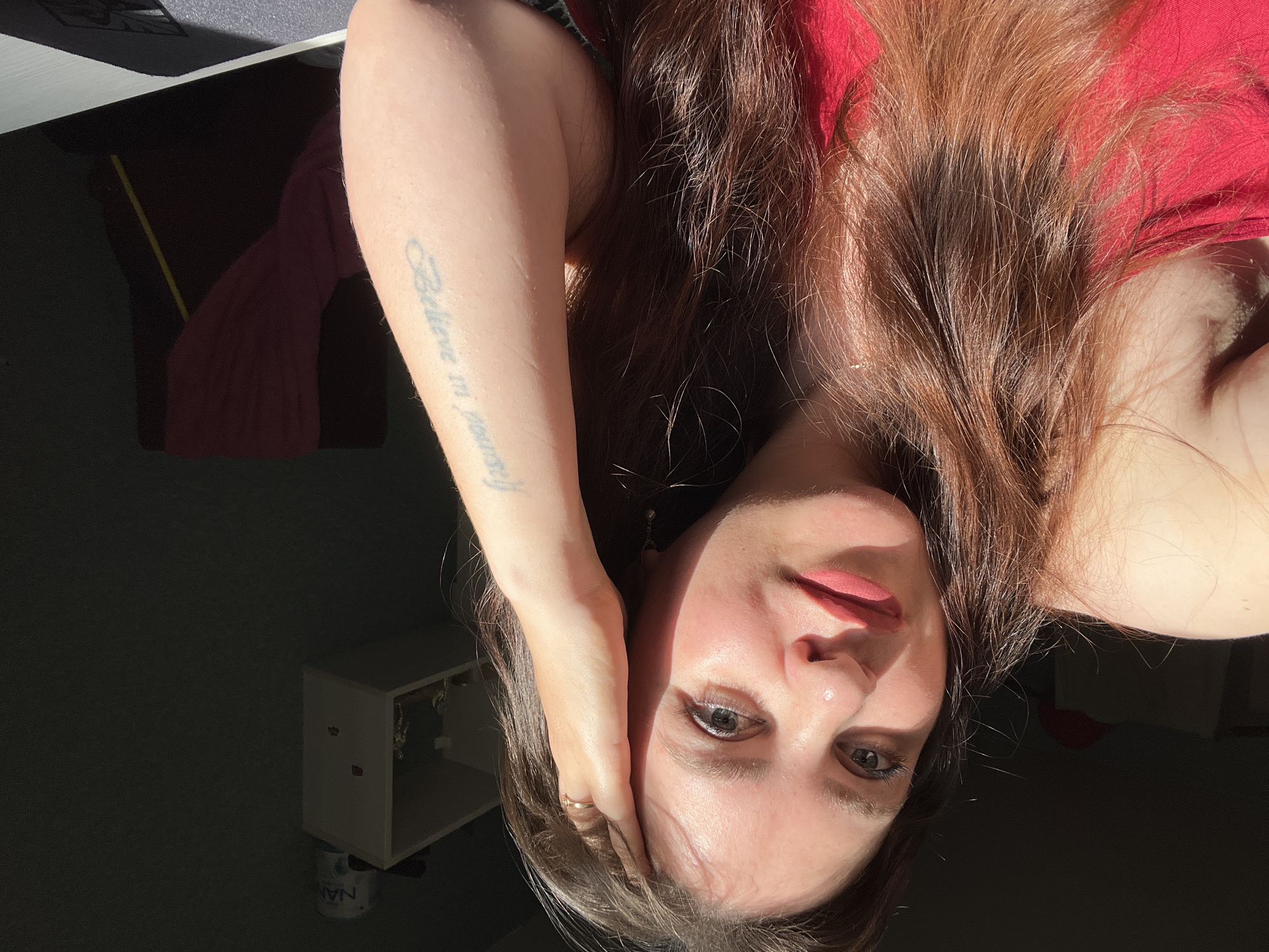 The image is a close-up selfie of a young woman with long, brown hair, looking directly at the camera. She is wearing a red top and has her hand on her head. A tattoo is visible on her left forearm, which reads "Believe in yourself." The background is dark, with a shelf on the right side of the frame containing a few objects, including a can labeled "NAN." Sunlight is streaming in from the left, casting a strong shadow across the woman's face and the scene. The setting appears to be indoors, possibly a room in Noviy, Russia, although the specific location within the city cannot be determined from the image alone.