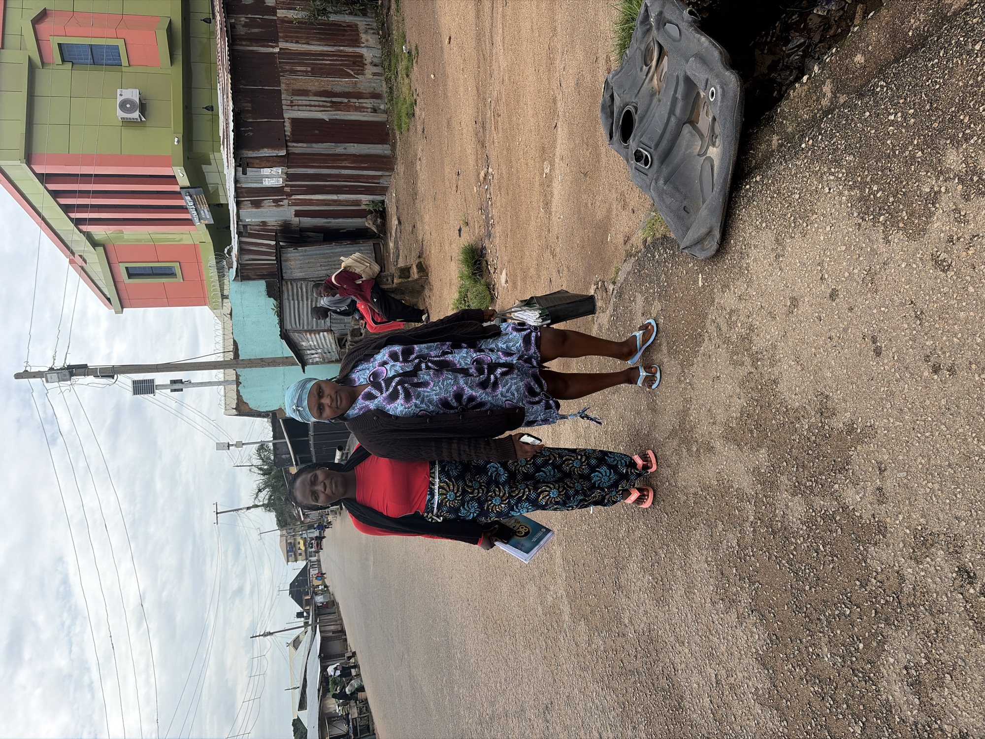 The image shows two women standing on a dusty, unpaved road in Jos, Nigeria. The woman on the left is wearing a red top and a patterned skirt, while the woman on the right is wearing a patterned dress and a dark cardigan. Both women are wearing flip-flops. They are standing in front of a row of buildings, some of which appear to be makeshift structures made of corrugated metal. In the background, there are more buildings, a few trees, and power lines. The sky is overcast. To the right of the women, there is a large piece of black plastic debris lying on the ground next to a ditch filled with trash. A modern, colorful building with signage is visible in the background.