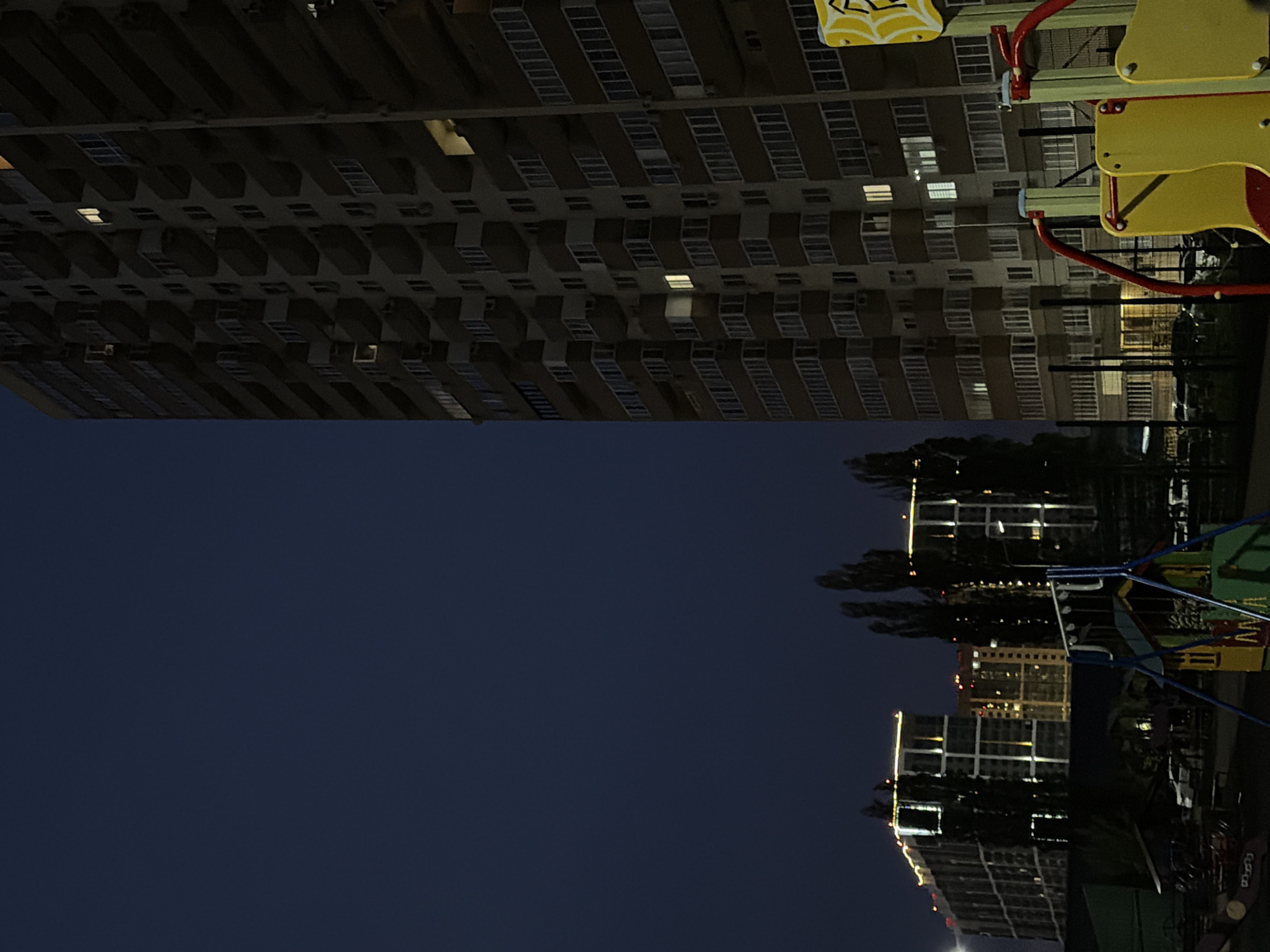 The image depicts an urban night scene, likely in Krasnodar, Russia, that has been rotated 90 degrees clockwise from its natural upright viewing position.
**Subjects:**
On the left side of the scene (which appears as the top of the image in its current rotation), a large, multi-story residential building stands tall. It features numerous windows and balconies, with several windows casting warm, yellow light. In the foreground, extending from the bottom right towards the center (appearing as the lower right in the current rotation), are colorful elements of a children's playground, including structures in yellow, red, blue, and green. The lower portion of the scene (appearing as the bottom of the image in its current rotation) features a dark, still body of water, reflecting the lights of other distant buildings and the dark silhouettes of trees, particularly on the right side.
**Activity or event taking place:**
No specific human activity or event is visible. The image captures a static, tranquil night view of a residential area.
**Notable details:**
The photograph was taken at night or in the late evening, evidenced by the dark sky and the prominent artificial lights illuminating the buildings and reflecting on the water. The sharp, clear reflections in the water suggest calm weather conditions. The overall color palette is dominated by dark blues and blacks, contrasted with bright points of light from the buildings.
**Visible text:**
There is no visible text in the image.