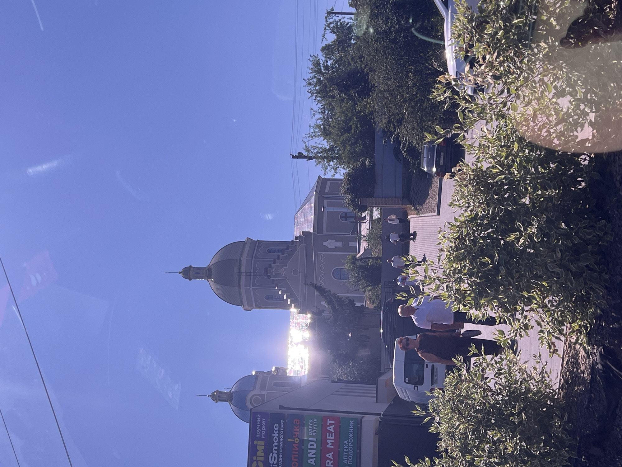In Kulykiv, Ukraine, on a sunny day, the facade of a building with two prominent domes is visible, with a bright sun glare obscuring part of the building's roof. In front of the building, a paved walkway leads upwards, with several people walking in different directions. Two individuals, a woman in a dark top and sunglasses and a man in a white polo shirt, are walking together towards the viewer. A white van is parked to their left. In the foreground, a row of green and white variegated bushes lines the walkway. Various signs are attached to the building on the left, including "Cimi", "iSmoke", "Копичка", "ANDI", and "ERA MEAT". The sky is clear and blue.