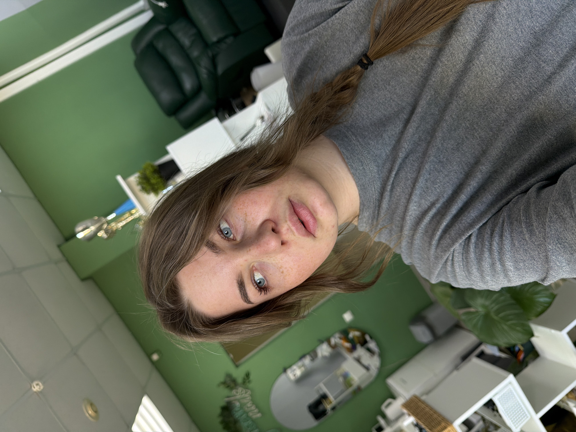 A selfie of a fair-skinned young woman with light brown hair partially pulled back in a ponytail, blue eyes, and freckles, wearing a gray long-sleeved shirt. She is indoors, possibly in a salon or office space. The background includes green walls, a white tile ceiling, a dark green leather chair, white cabinets or shelving, a mirror, plants, and other salon/office equipment. The location is in the city of Chaikovsky, Russia.