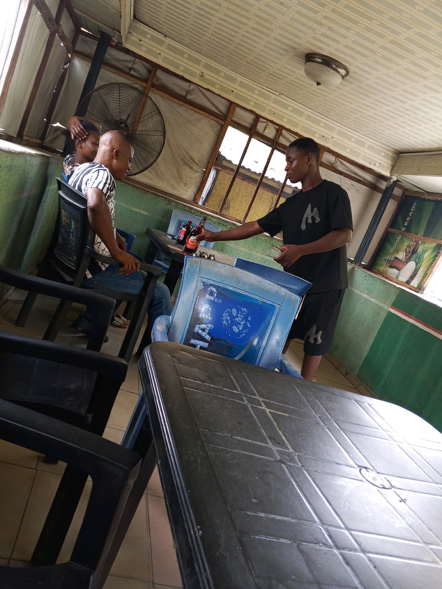In Zango-ndaji, Nigeria, at a covered outdoor seating area, two young men are engaged at a table. One man, wearing a black t-shirt with a white logo and black shorts, stands while holding a red bottle and a phone. He appears to be interacting with the other man who is seated. The seated man, wearing a black and white striped t-shirt and blue jeans, looks towards the standing man. On the table between them are two more red bottles, a pair of glasses, and what appears to be a small metal tin. The environment is sparsely furnished with black plastic chairs and tables, one of which is partially visible in the foreground. A large industrial fan is mounted on the wall behind the seated man, and posters are visible on the back wall. The ceiling is made of corrugated metal panels.