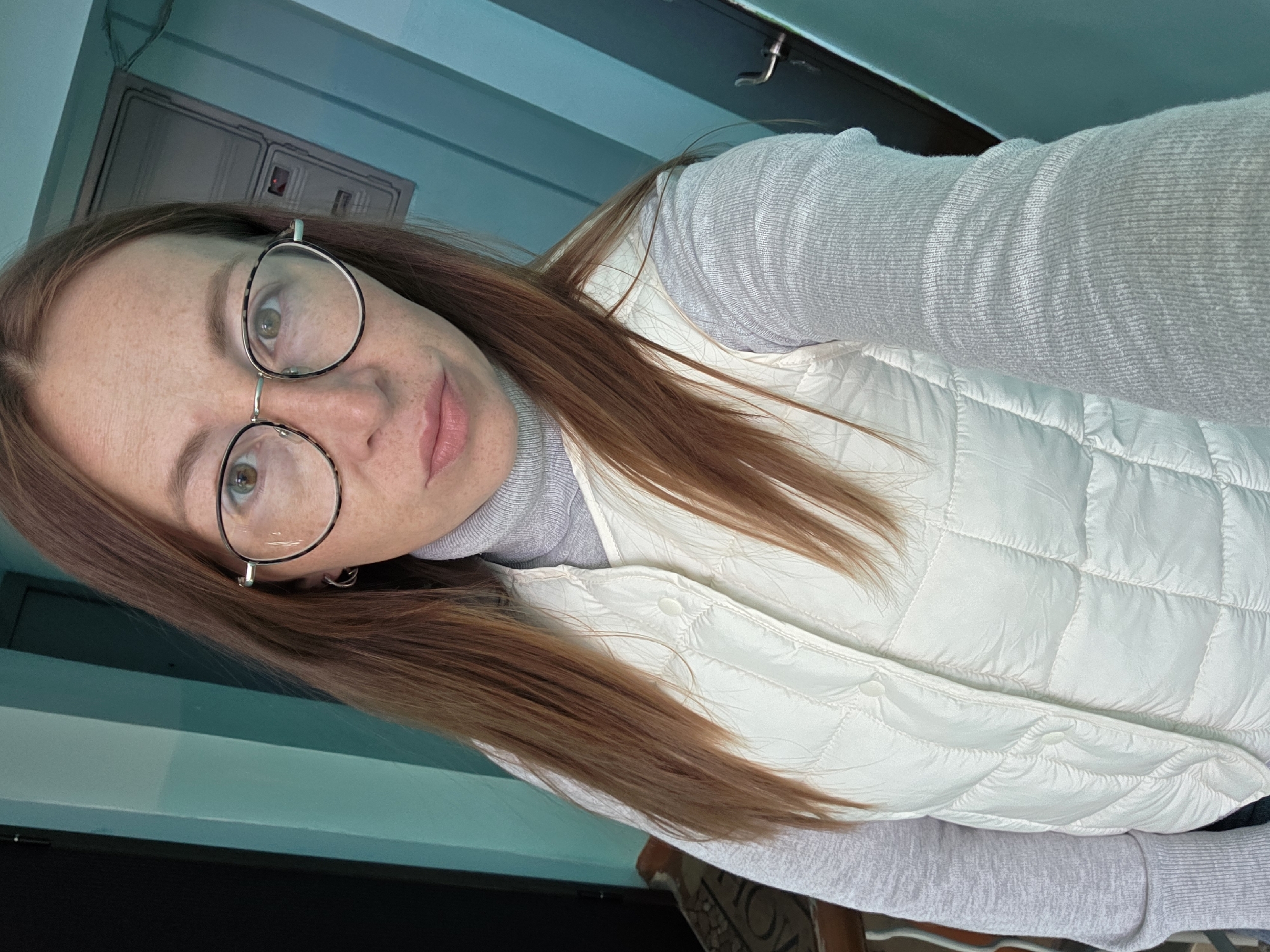 A woman with long reddish-brown hair, fair skin, and freckles is wearing round-framed eyeglasses. She has on a light gray turtleneck sweater and a white quilted vest. The background appears to be a room with blue walls and a black ceiling area. The location is not explicitly stated in the image itself to be Chaikovsky, Russia, and cannot be definitively determined from the visual information provided.