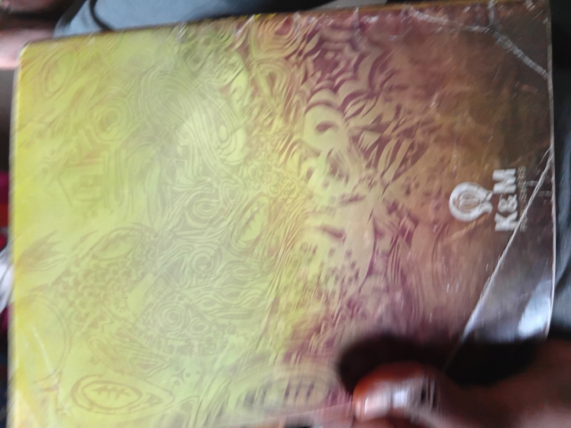 A person's hand is visible on the left side of the image, holding a book or binder. The cover of the book is yellow with a patterned design. In the lower center of the book cover, there is a logo with the letters "K&M" and the word "PUBLISHERS" underneath. The image appears to be taken indoors, possibly in a home or office setting. The specific location in Nkpor, Nigeria, cannot be determined from the provided image.