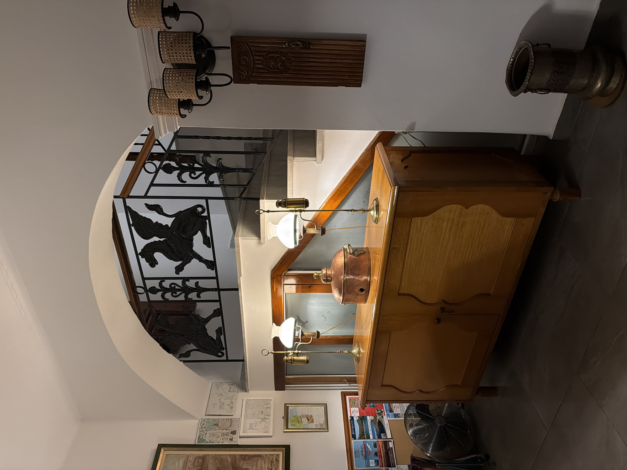 An interior shot taken from a slightly low angle showcases a section of a room near a staircase in what appears to be a building in Hydra, Greece. The staircase is adorned with a wrought iron railing featuring stylized griffins or similar mythical creatures.
To the right of the frame, a light-colored wooden cabinet with doors stands, with two antique-style oil lamps placed on top. One lamp is brass-colored, while the other one features a copper container. Above the cabinet, mounted on the wall, are two light fixtures; one is a multi-lamp wall sconce and the other a carved wooden panel.
Below the staircase, several framed pictures hang on the wall. In the lower part of the image, fragments of additional decor and furniture, including a fan, are visible. The floor is tiled with gray, elongated tiles. The color palette is warm, with the wooden furniture and soft lighting contrasting against the white walls. The environment suggests a historic building with a blend of traditional and classic elements.