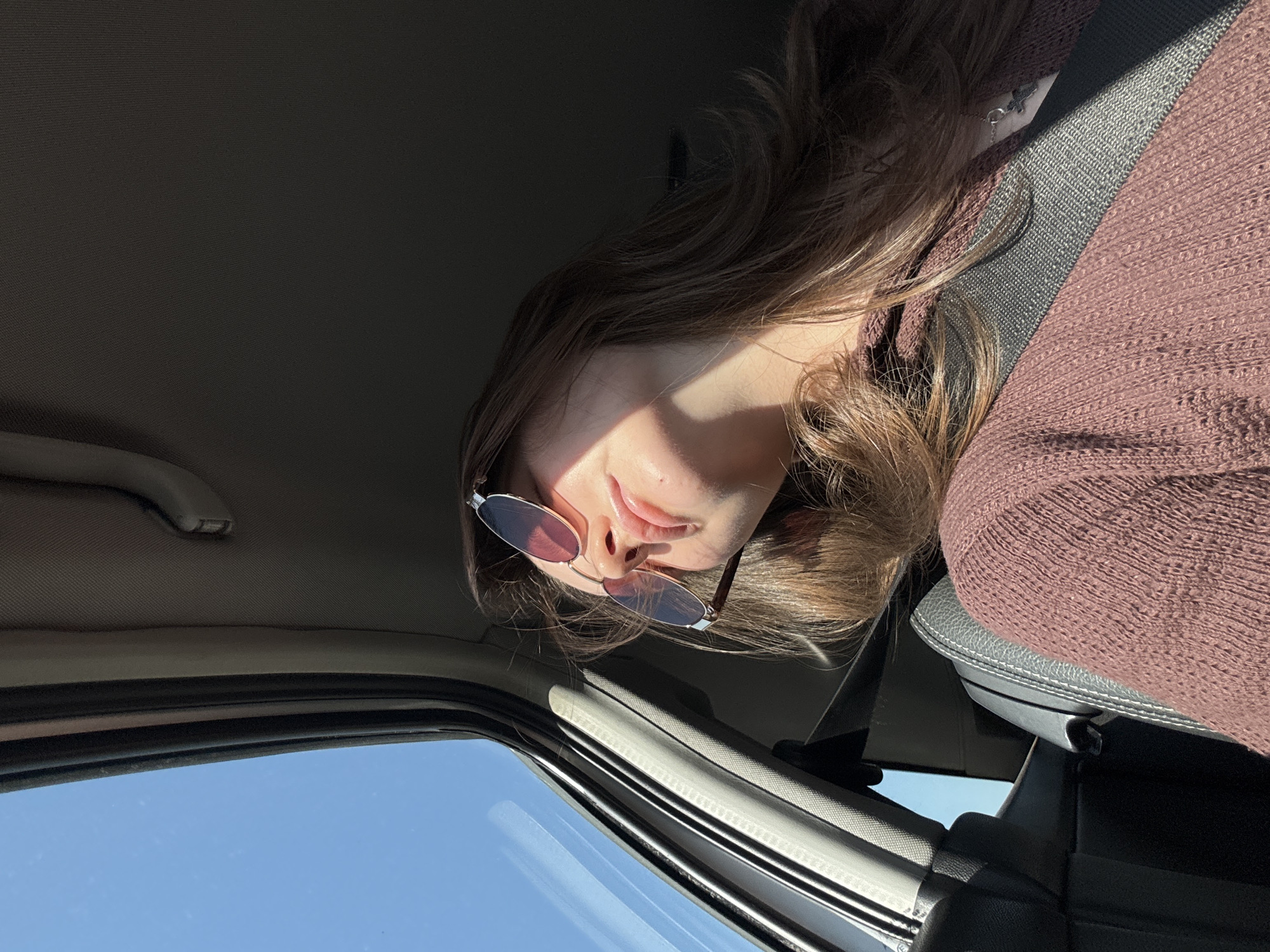 A medium shot, taken from a low angle inside a car, shows a young woman wearing sunglasses and a mauve-colored sweater. She is seated in the passenger seat, with a partially visible seatbelt. The view through the car window shows a bright blue sky. The location information suggests this scene is taking place in Шумиха, Russia.