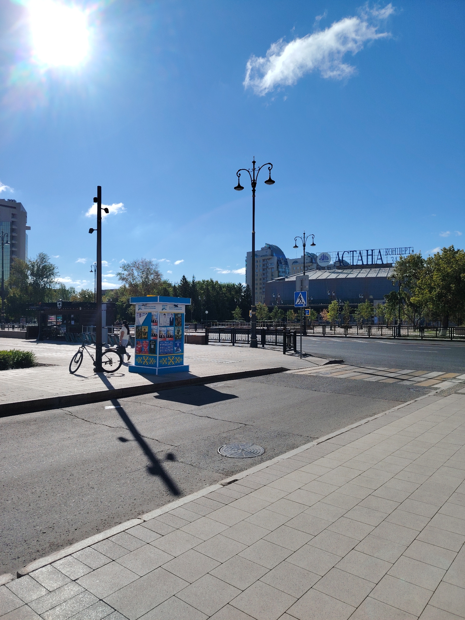 A bright, sunny day in Astana, Kazakhstan, features a wide paved area with a sidewalk made of light-colored rectangular tiles. A street runs parallel to the sidewalk, with a pedestrian crossing marked by white stripes. A black bicycle is parked near a lamppost on the sidewalk, casting a long shadow. Adjacent to the bicycle is a blue and white kiosk displaying posters. In the background, several modern buildings rise, including a large building with "АСТАНА КОНЦЕРТ ЗАЛ" (Astana Concert Hall) emblazoned on its facade. Trees line the street, and a pedestrian signal with a walking person icon is visible. The sky is a clear blue with a few wispy white clouds, and the sun shines brightly, creating lens flare.
