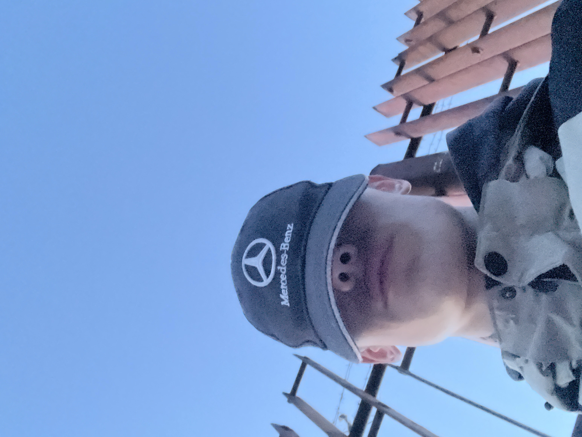 The image shows a low-angle, close-up selfie of a young person wearing a dark baseball cap with a white Mercedes-Benz logo and text. The brim of the cap covers the person's eyes, obscuring their face, but their nose and mouth are visible. They are wearing a camouflage jacket over a dark hooded garment. The background consists of a clear blue sky and a section of a rusty metal fence. The setting appears to be outdoors, possibly in a public area or park. The photograph was taken in the city of Noviy, Russia.