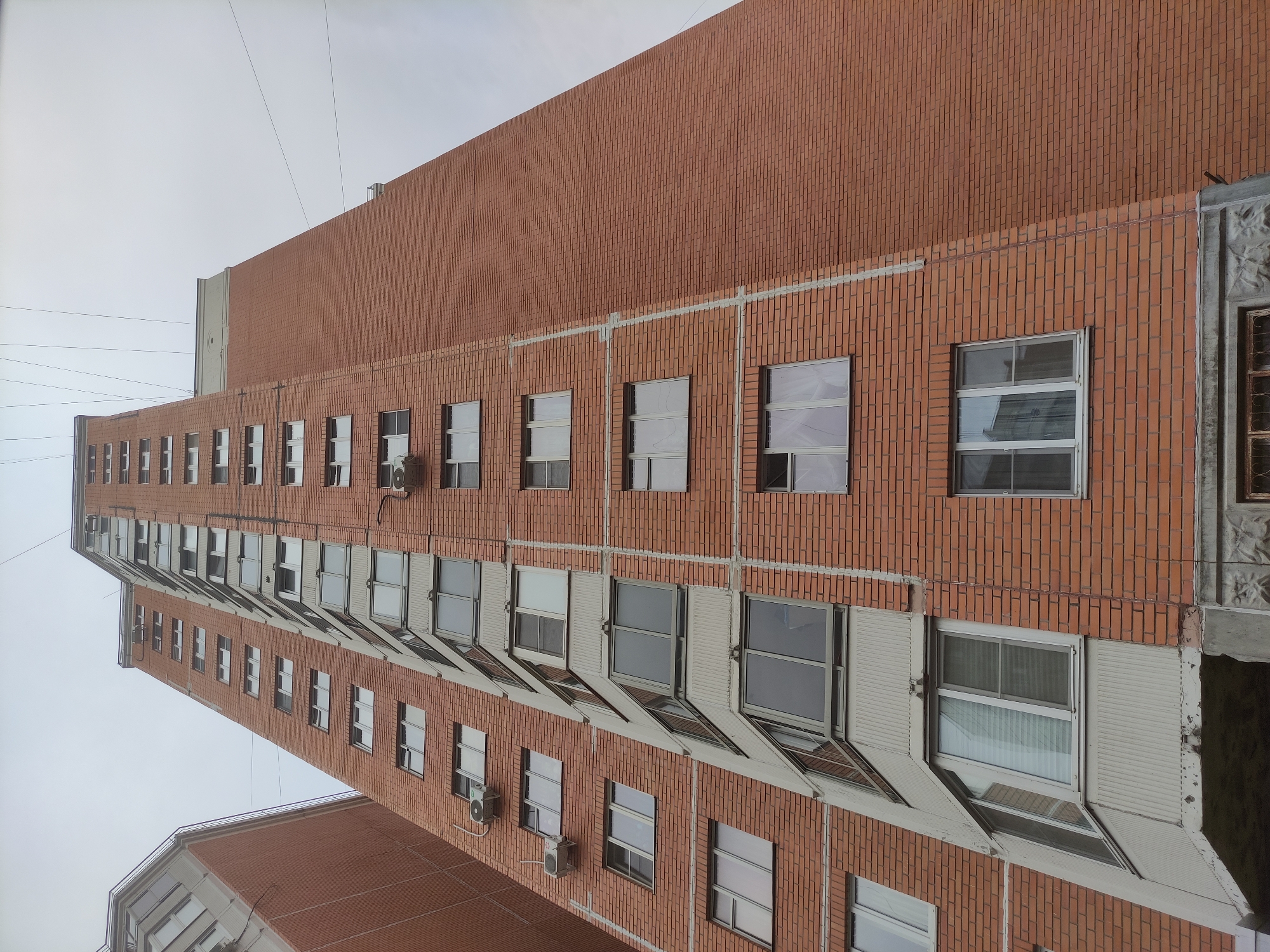 The image shows the facade of a multi-story brick building in Khimki, Russia. The building is primarily constructed of red bricks. Windows are present on each floor, and some have external air conditioning units installed. The sky is overcast.