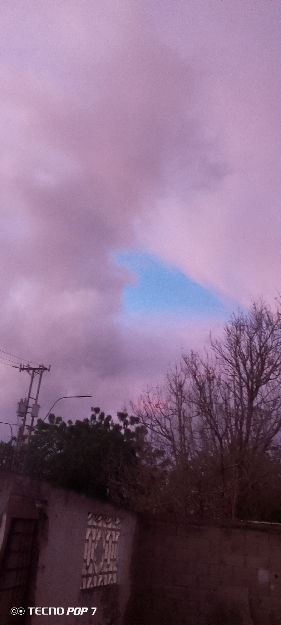 The image captures an outdoor scene at what appears to be either dawn or dusk, characterized by a dramatic and colorful sky. The dominant feature is the expansive sky, filled with soft, swirling clouds in varying shades of purple and pink. A distinct, irregular patch of bright blue sky is visible near the center-right, breaking through the cloud cover.
In the lower portion of the image, elements of an urban or semi-urban environment are present. On the left, a utility pole with multiple electrical wires extends upwards against the cloudy backdrop. Below it, dark green tree foliage is visible, partially obscuring a building structure with a window covered by security bars. To the right, the bare, intricate branches of deciduous trees stand silhouetted against the vibrant sky. Another part of a building or wall made of bricks is visible in the bottom right corner.
No people are visible, and no specific activity is taking place; the image presents a still landscape or street view. The atmospheric lighting, characterized by the purple and pink hues, suggests a transitional time of day or unique weather conditions. The scene is consistent with a residential or suburban area in Altagracia, Venezuela. In the bottom left corner, the text "OO TECNO POP 7" is visible, likely a camera watermark.
