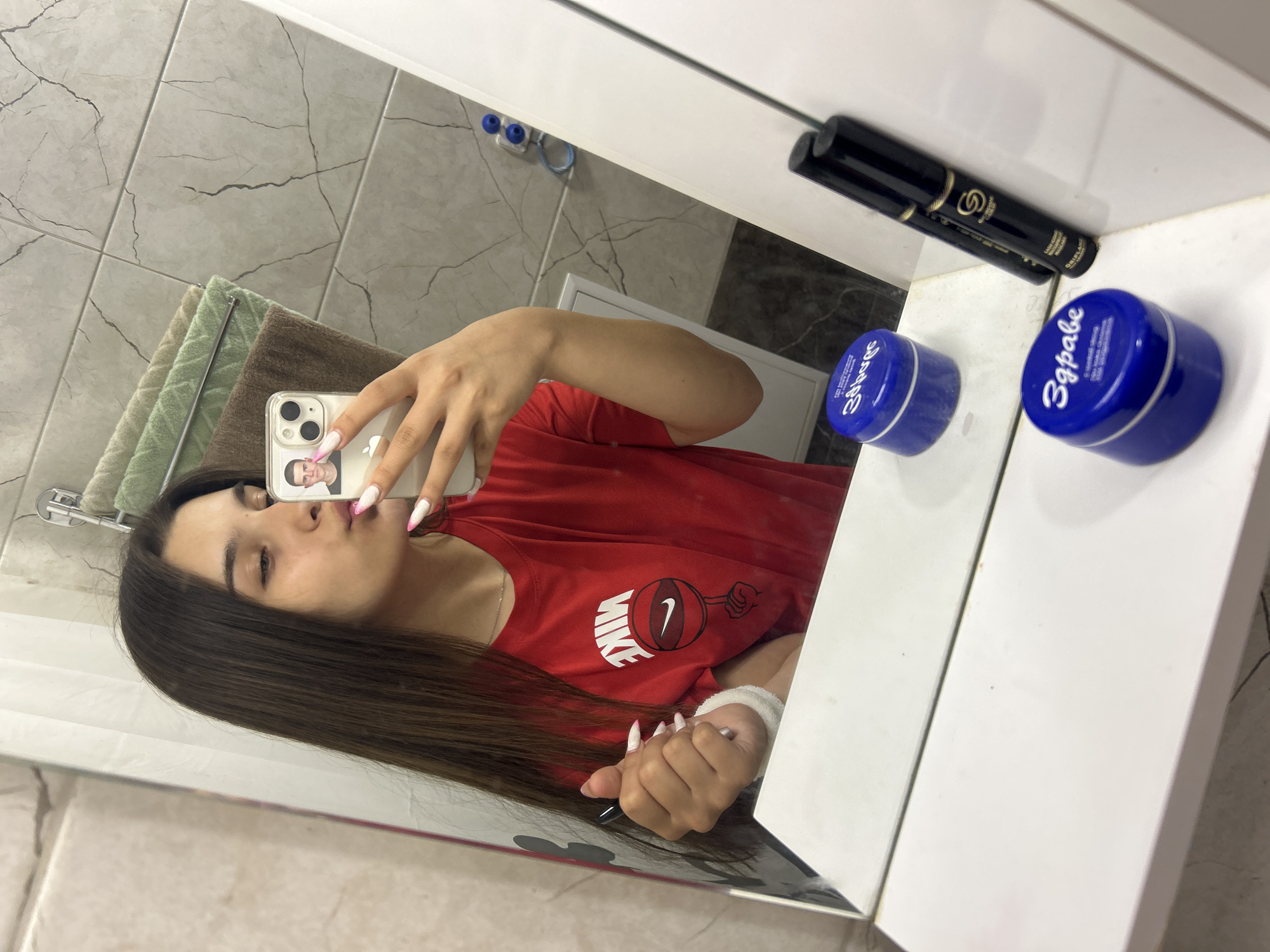 A young woman with long brown hair is taking a selfie in a bathroom mirror. She is wearing a red t-shirt with a Nike logo. She is holding a phone in her right hand and has a pen in her left hand. There are two blue cosmetic jars and a mascara tube on the counter in front of the mirror. The bathroom has tiled walls and a shelf with towels. The scene is likely in the city of Shumen, Bulgaria, given the text on the cosmetic jars, which appears to be in Bulgarian.