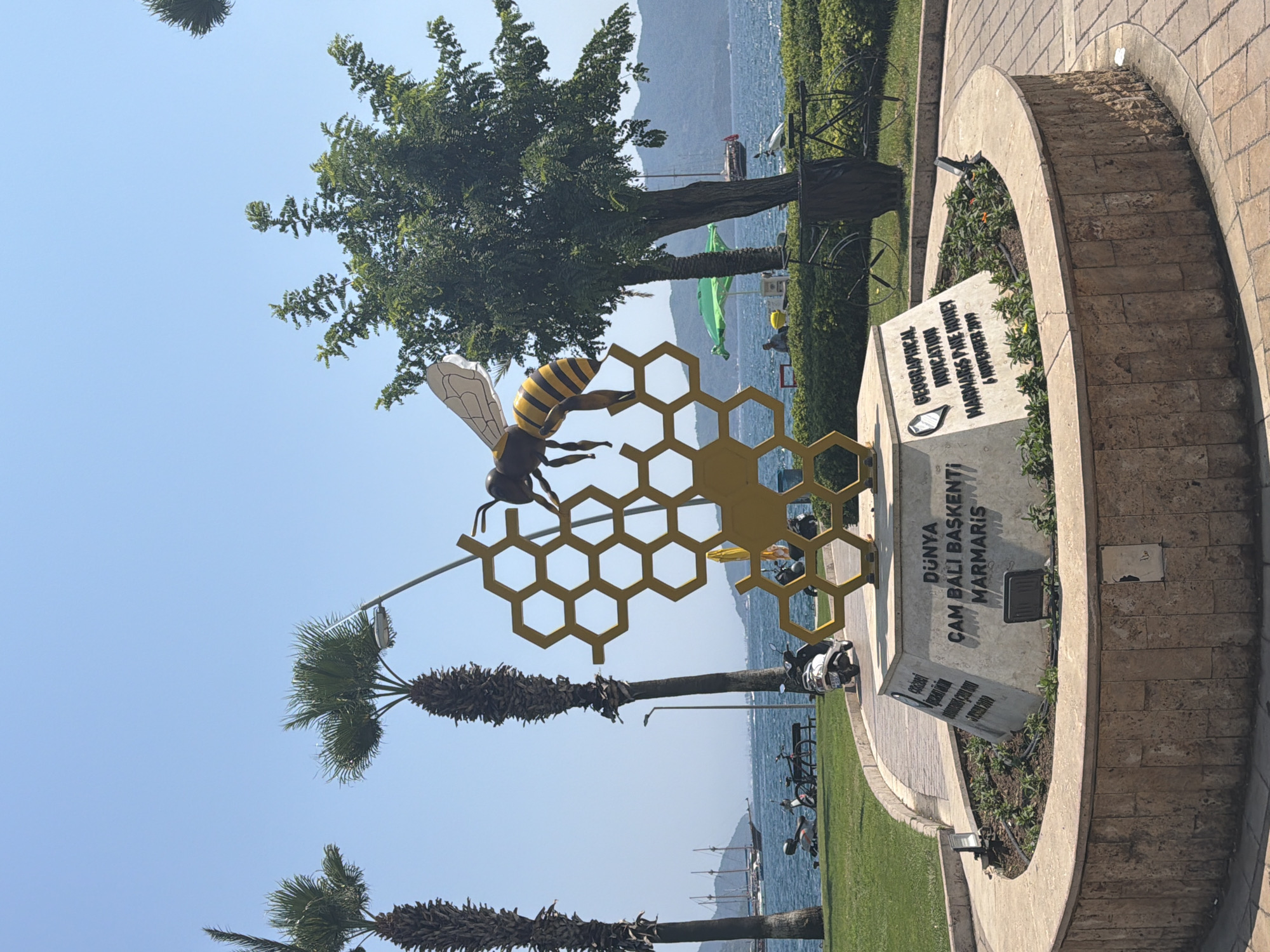 A sculpture of a bee on a honeycomb is located in the city of Marmaris, Turkey, near a waterfront. The sculpture stands on a circular stone base inscribed with text in Turkish and English. The Turkish inscription reads "DÜNYA ÇAM BALI BAŞKENTİ MARMARİS" (World Pine Honey Capital Marmaris), and the English inscription reads "GEOGRAPHICAL INDICATION MARMARİS PINE HONEY 6 NOVEMBER 2019." Several palm trees and a large leafy tree are visible in the background, along with a body of water, boats, and a distant mountain range. A few scooters and bicycles are parked on the grass in the middle ground. The sky is clear and blue.