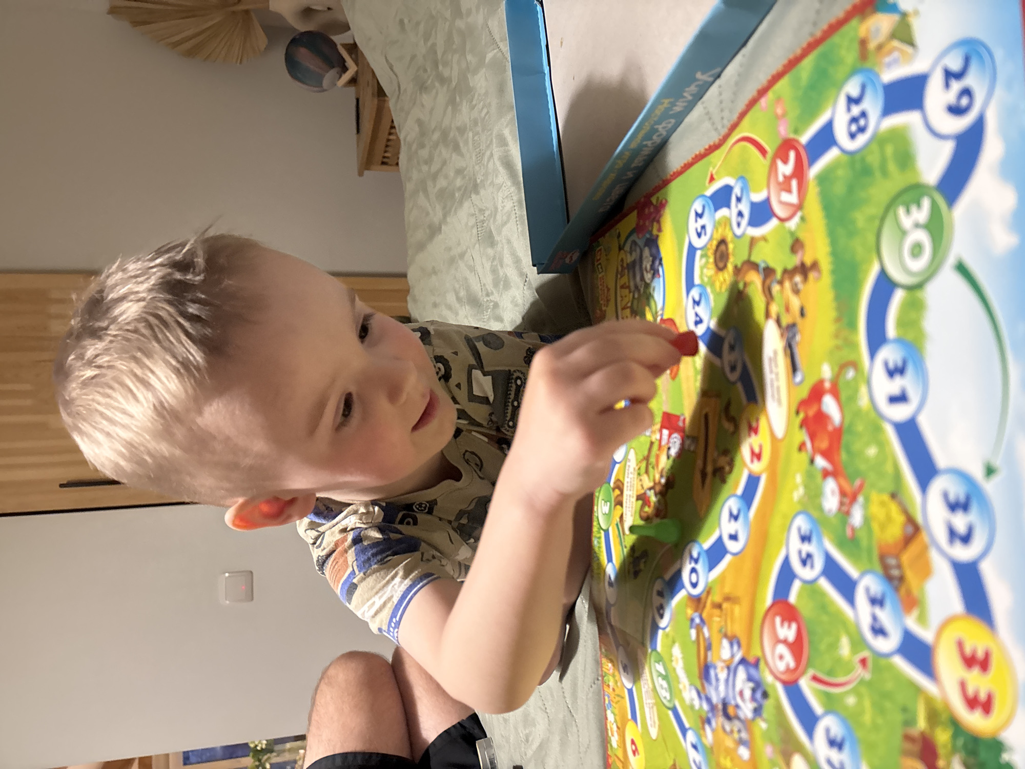 The image shows a young, light-skinned child with short blonde hair playing a board game. The game board is large and colorful, featuring numbered spaces and cartoon illustrations, with a blue game box nearby. The child is holding a red game piece. The background includes a portion of a bed, a light switch on a wall, and a leg covered in black shorts or trousers. The location appears to be indoors, potentially in a residence, though without more context, it's impossible to confirm that this photo was taken in Chaikovsky, Russia.