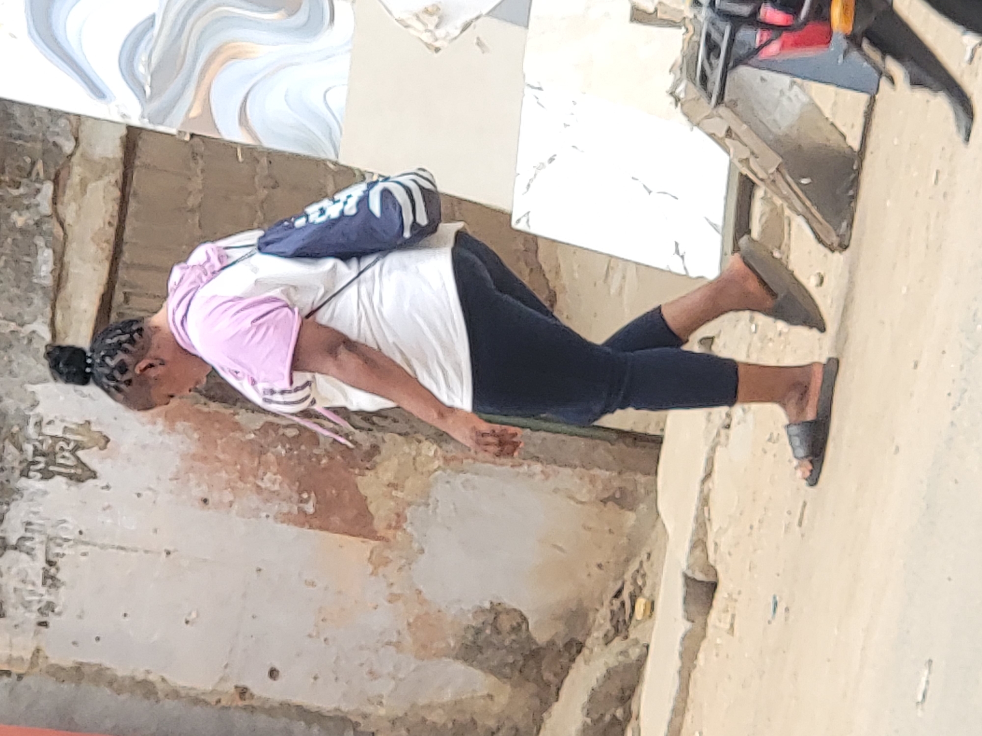 A woman stands in a dilapidated building in Maiburiji, Nigeria. She has dark hair pulled back in a bun and is wearing a white t-shirt with a pink and white striped sweater over it, dark blue leggings, and black sandals. She is also wearing a dark blue backpack. The building is in disrepair, with crumbling walls and exposed beams. A piece of art hangs on the wall near the ceiling. Debris is visible on the floor.