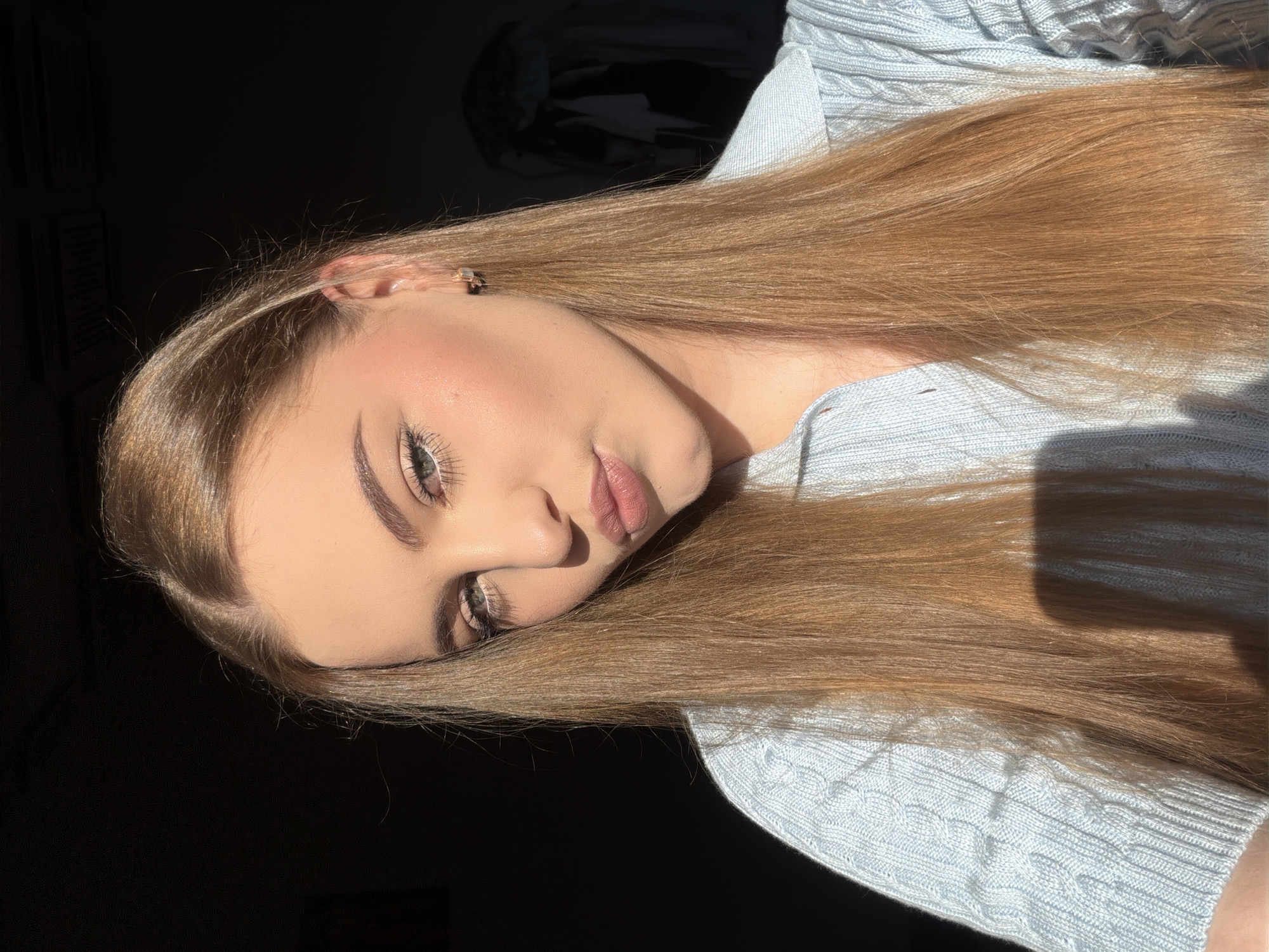 A young woman with long, straight, light brown hair is captured in a close-up, selfie-style shot. She is wearing a light blue, cable-knit sweater with a collared neckline. Her makeup includes winged eyeliner and lip gloss. She is looking towards the camera with her lips pursed. The lighting suggests she is near a window, with sunlight illuminating her face and casting a shadow to her left. The background is dark, with some indistinct shapes visible on the right, possibly clothing on a rack. There are no visible landmarks or cues that would indicate a specific location within the city of Dnipro, Ukraine.