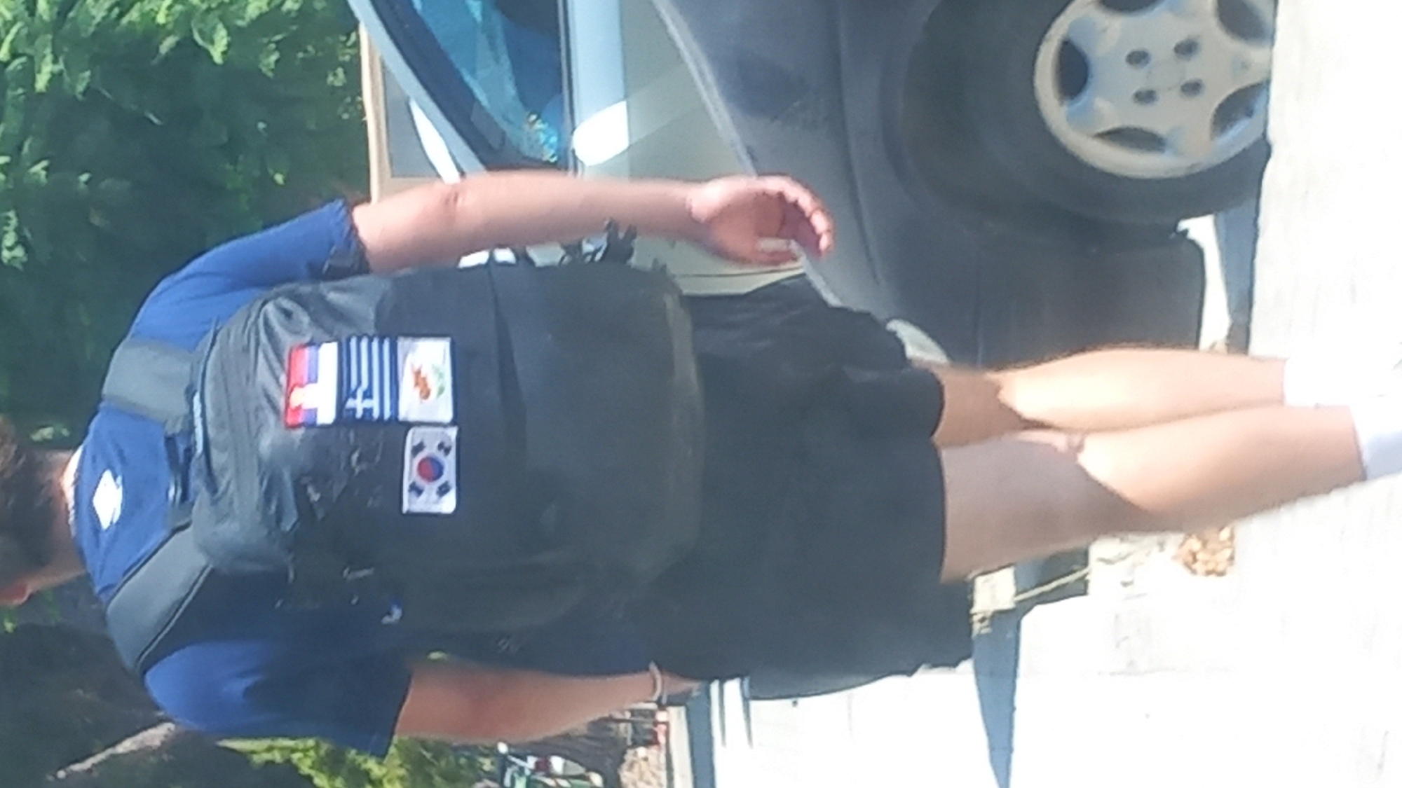 A person wearing a blue t-shirt and black shorts walks away from the camera, carrying a black backpack adorned with several flag patches. The patches appear to represent South Korea, Greece, and potentially Cyprus and another nation. The person is on a sidewalk next to a parked car, with trees and foliage in the background. The scene is outdoors in daylight. The image appears to have been taken in the city of Kos, Greece.