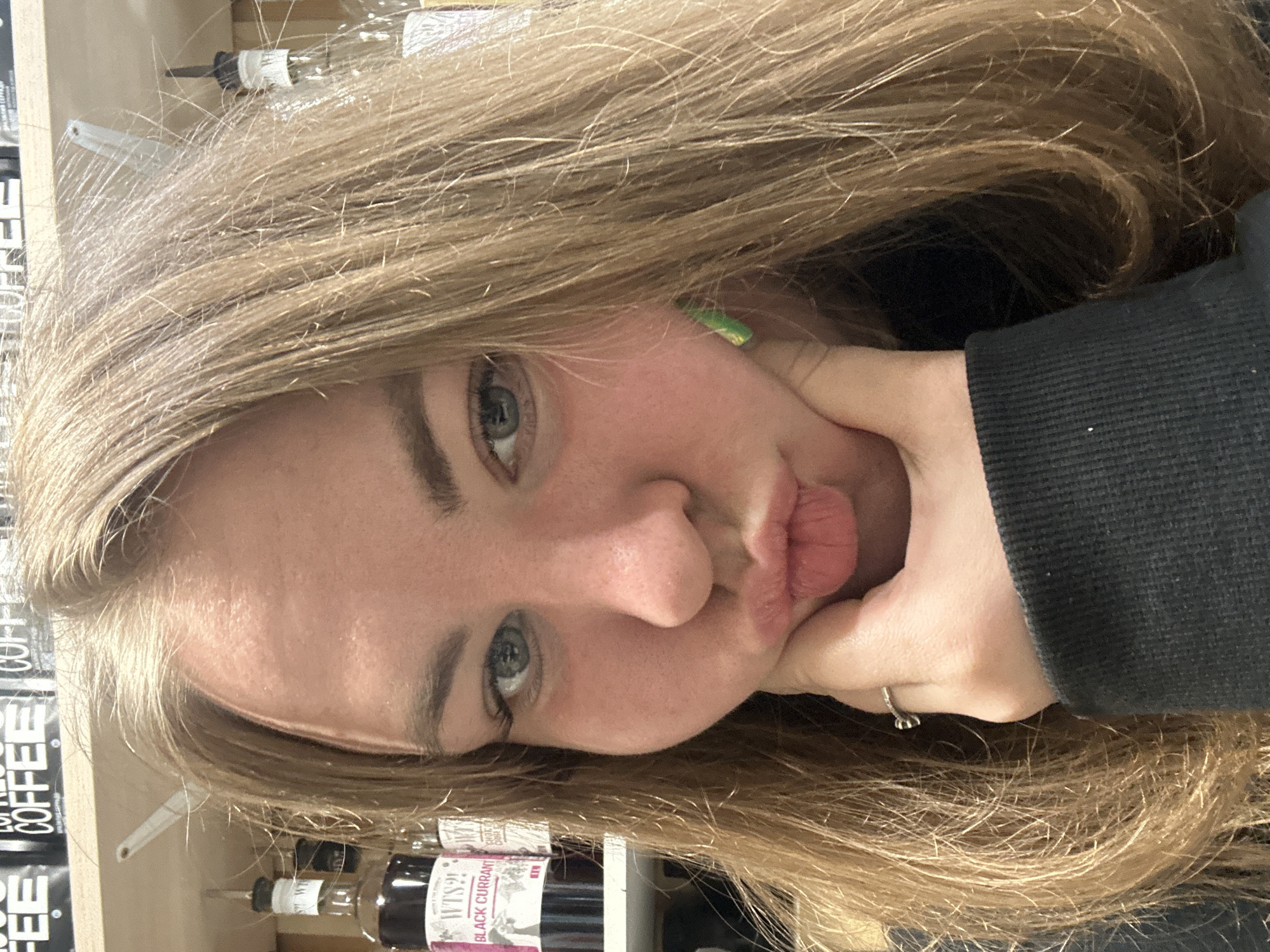 A close-up portrait shows a young person with long, light brown hair, tilted to the right. The individual's face is prominent, featuring light-colored eyes (the left eye is clearly visible) and dark eyebrows. Their lips are puckered into a "kissing face" or pout. A hand, likely the person's right hand, cups the chin and lower cheek. A silver-colored ring with a distinctive shape is visible on the ring finger. The person is wearing a dark, possibly black, long-sleeved garment, partially seen on the right side of the frame. A small, bright green object or accessory is visible near the person's right ear.
The background consists of light-colored shelving, populated with various bottles and containers. Some items appear to be coffee-related products, with the word "COFFEE" visible on one package. Another clear bottle on the bottom shelf has a label partially reading "WTS?!" and "BLACK CURRANT," possibly indicating a syrup or flavoring. Another bottle nearby has a dropper top. The setting suggests an indoor environment, possibly a kitchen, pantry, or a retail space. The overall impression is a casual and playful moment captured in a close-up shot.