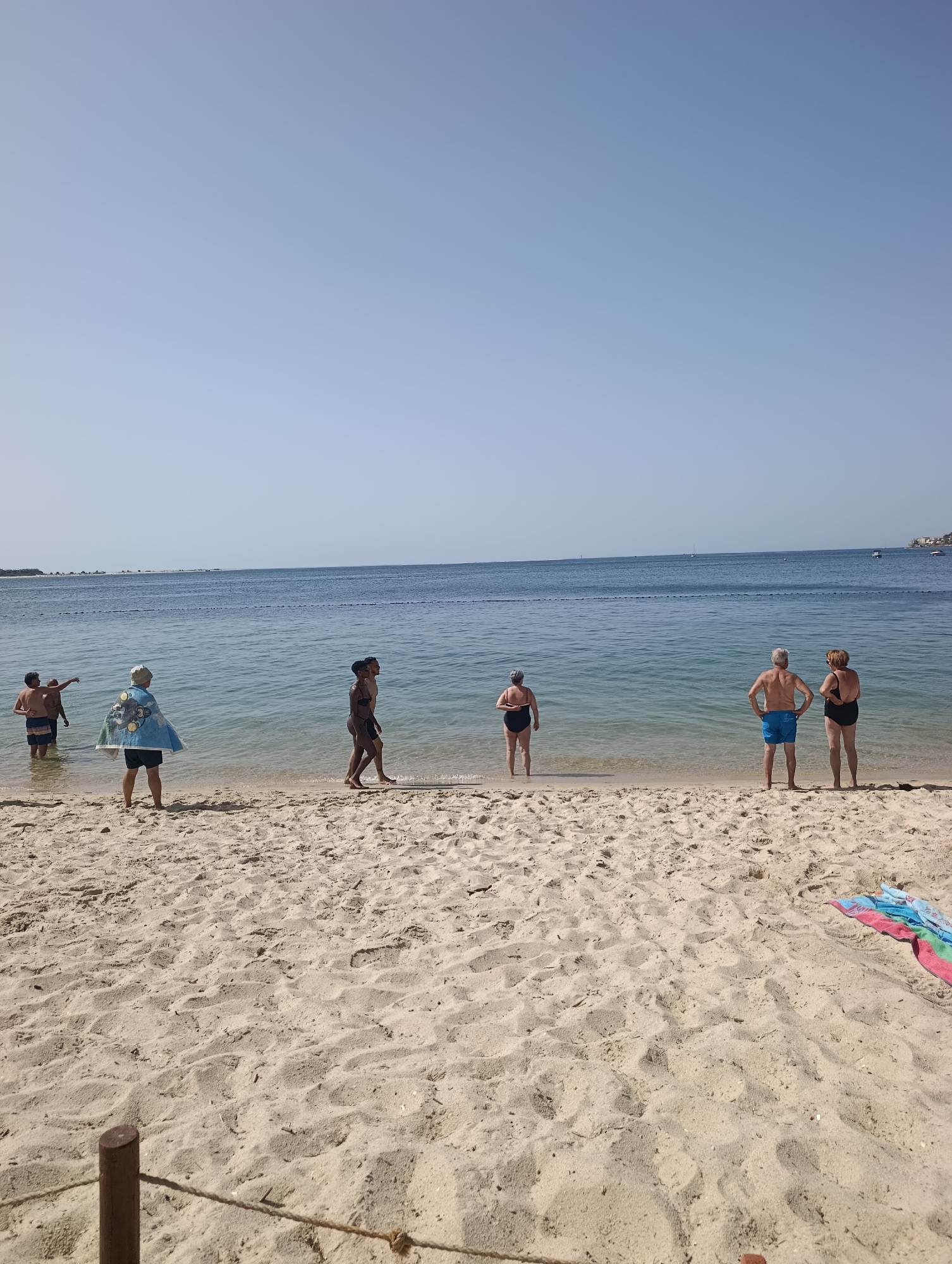 The image depicts a beach scene with several people near the water's edge. The setting appears to be a sunny day with a clear blue sky. The sand is light in color and appears undisturbed except for footprints. The water is calm and a deep blue, with a rope barrier visible in the distance across the water.
On the left side of the image, two men are standing in the shallow water. One of them is pointing towards something out of frame. Between them and the water's edge, a person wearing a colorful blanket over their shoulders stands facing away from the camera. To the right of this person, two individuals are walking along the shoreline, facing left. Further to the right, a man and a woman are standing on the sand, facing the water. On the far right, near the water, a woman in a dark swimsuit is standing. A colorful towel is spread on the sand in the lower right corner. In the background, a distant coastline with some structures is visible.
The specific location in Setúbal, Portugal is not identifiable from the image alone. However, the scene is consistent with a typical beach found along the Portuguese coast.