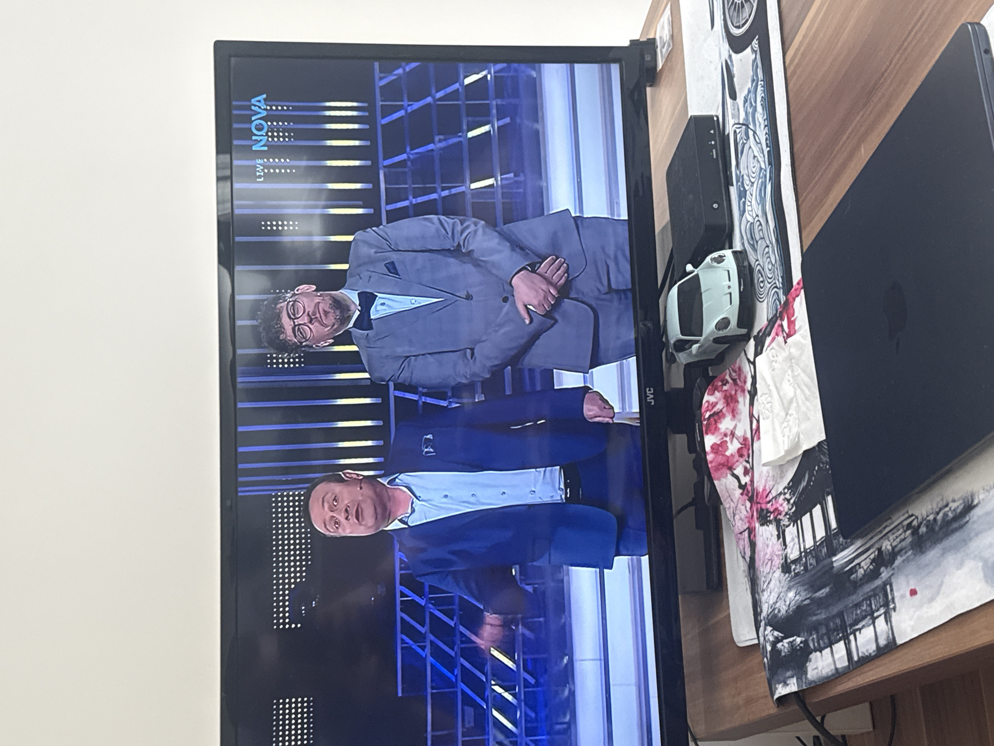 A television screen displays two men in suits on what appears to be a set for a TV show. The television is positioned on a wooden surface next to a laptop, a black set-top box, a toy car, and a mousepad with a floral and architectural design. The scene appears to be indoors, against a white wall. There is no direct indication of the city of Sofia, Bulgaria.