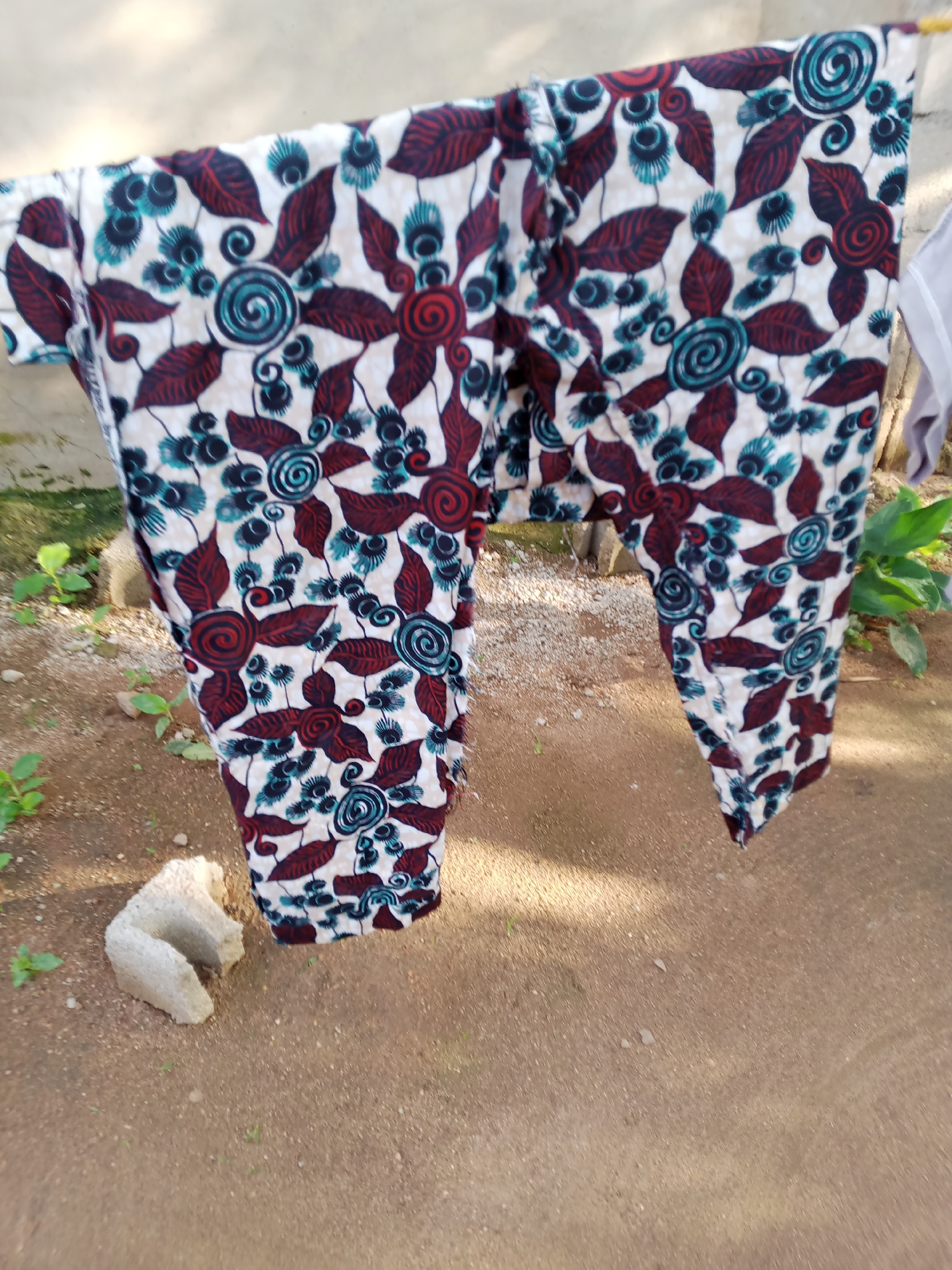 A pair of colorful African print trousers are hanging on a clothesline outdoors. The fabric features a pattern of white, maroon, and teal. The background is a mix of dirt, gravel, and some sparse green vegetation. To the left of the trousers, there is a light-colored, irregular-shaped object on the ground. In the distance, a concrete wall is visible. The overall scene suggests a domestic setting in Jauro Yinu, Nigeria.
