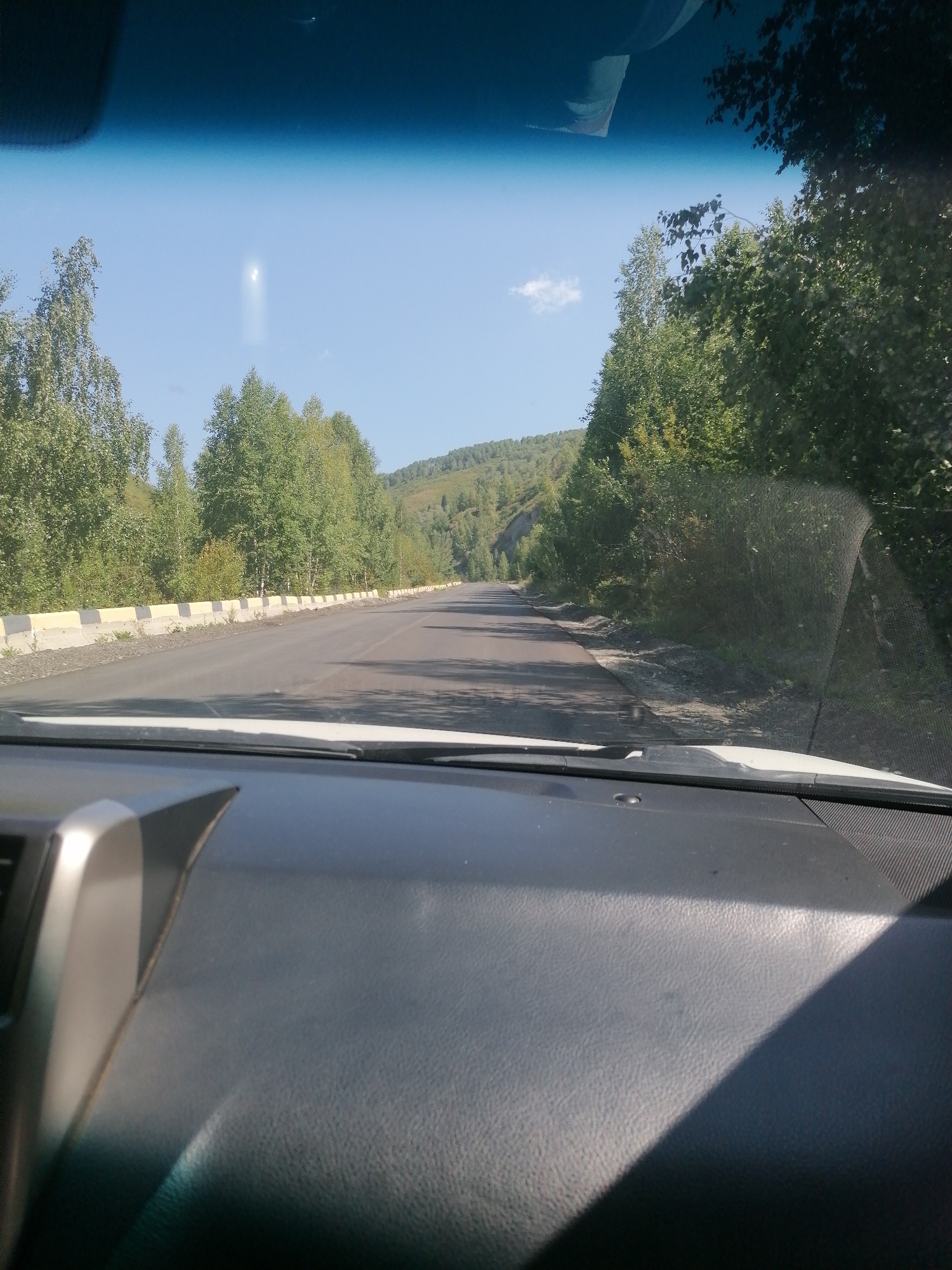 The image shows a view from inside a vehicle, looking out through the windshield. The road is paved and appears to be a two-lane highway. On the right side of the road, there is a curb with yellow and white concrete barriers. Beyond the barriers, there are numerous trees and vegetation that cover the slopes of rolling hills. The sky is clear and blue with a small cloud visible in the distance. There is a bright light, possibly the sun, visible in the sky. The dashboard of the vehicle is visible in the foreground. The scene is set in a natural, rural environment, likely a mountainous or hilly region. The location is identified as Tarkhanka Rural District, Kazakhstan.