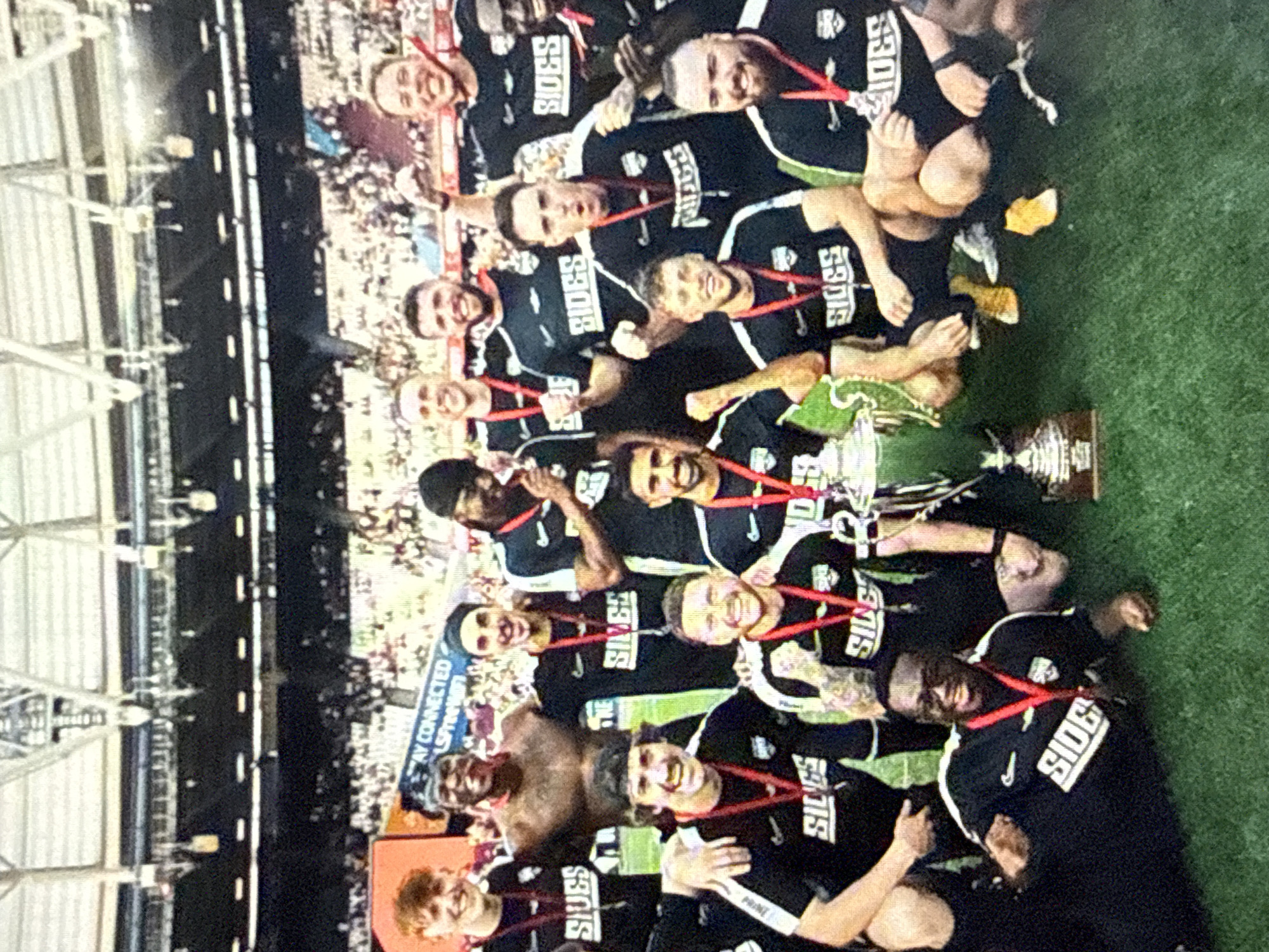 A group of soccer players, wearing black jerseys with "SIDES" written on them, are gathered on a grass field, posing for a picture while holding a trophy. They are wearing medals around their necks. Behind them is a crowd of people and part of a stadium structure. The location is in London, United Kingdom, based on the stadium's design.