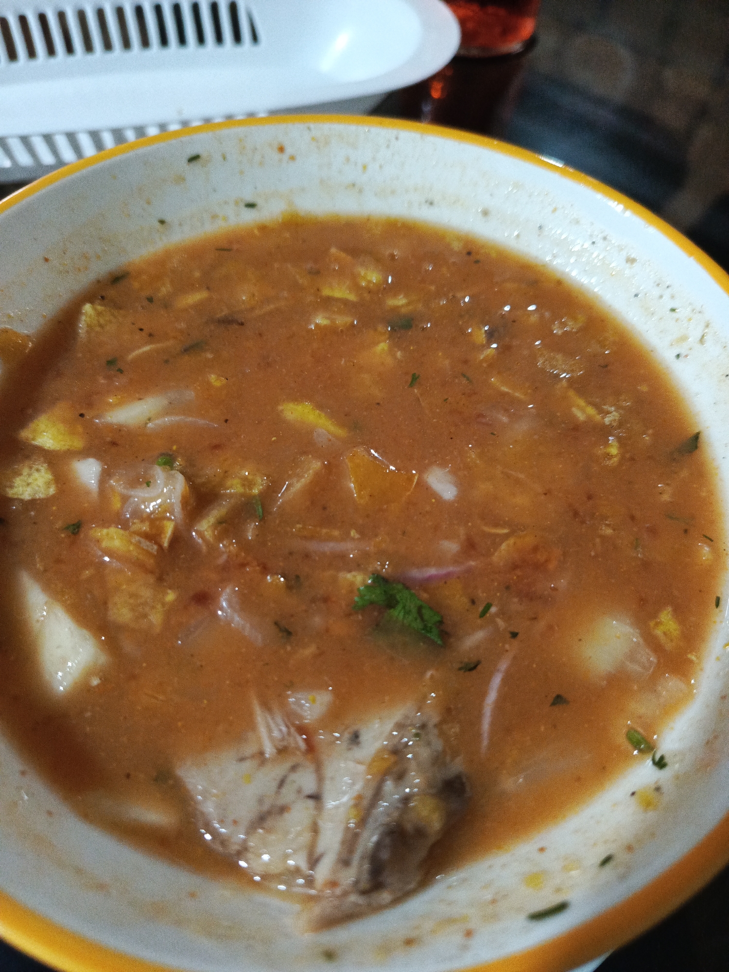 The image shows a close-up of a bowl of soup with visible chunks of food, including what appears to be meat and vegetables. The soup is a reddish-orange color. The bowl has a yellow rim. In the background, there is a white object with parallel lines, possibly a rack or a basket, and a dark surface that might be a table. A blurry red glass is also visible in the upper right corner. The scene appears to be indoors, likely a restaurant or home setting in Guayaquil, Ecuador. There are no people or discernible actions taking place.