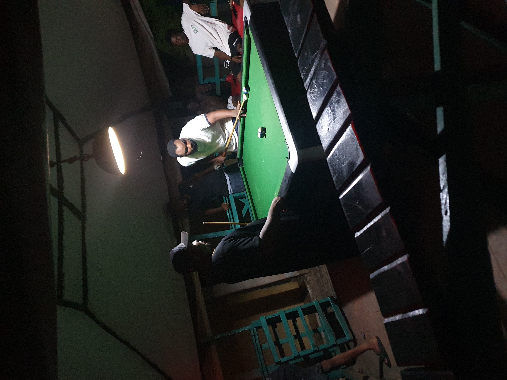 People are playing billiards in a dimly lit indoor space, with an overhead lamp illuminating the pool table. The location appears to be in Lokoja, Nigeria, as the lighting suggests. There are four people visible around the pool table. One person is actively playing billiards, while the other three people are watching. The environment includes a table, walls, and supports.