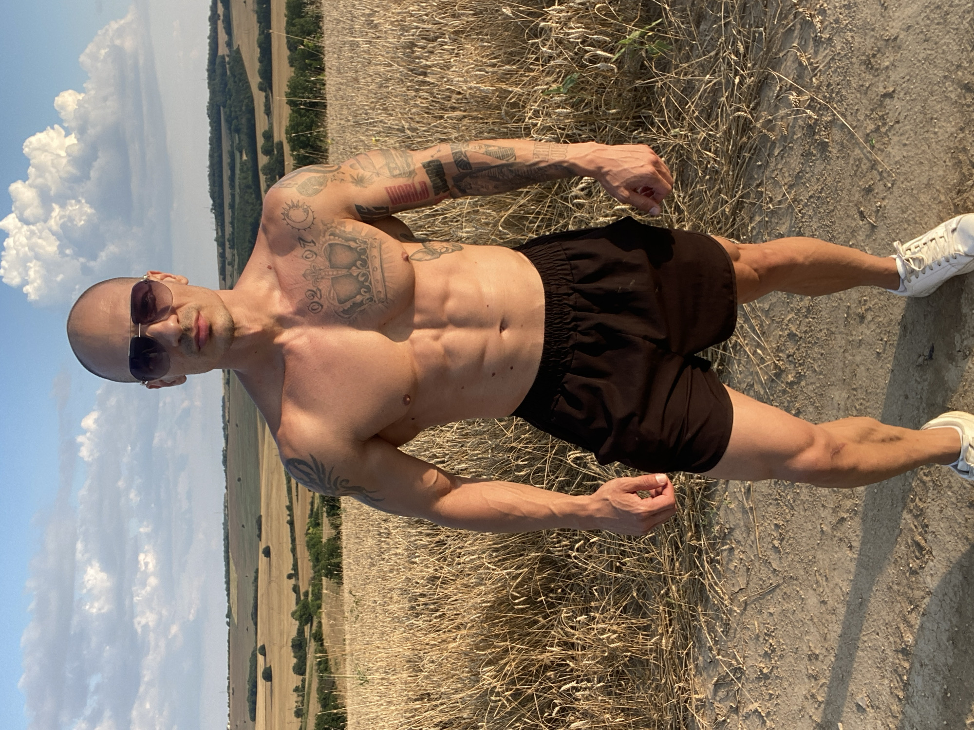 An adult male with a shaved head, sunglasses, and muscular physique stands in a field of dry wheat. He is wearing black shorts and white sneakers. The man has tattoos on his chest, arms, and shoulders. In the background, there are rolling hills with sparse trees and a partly cloudy sky. The image appears to have been taken in the rural outskirts of Pleven, Bulgaria.