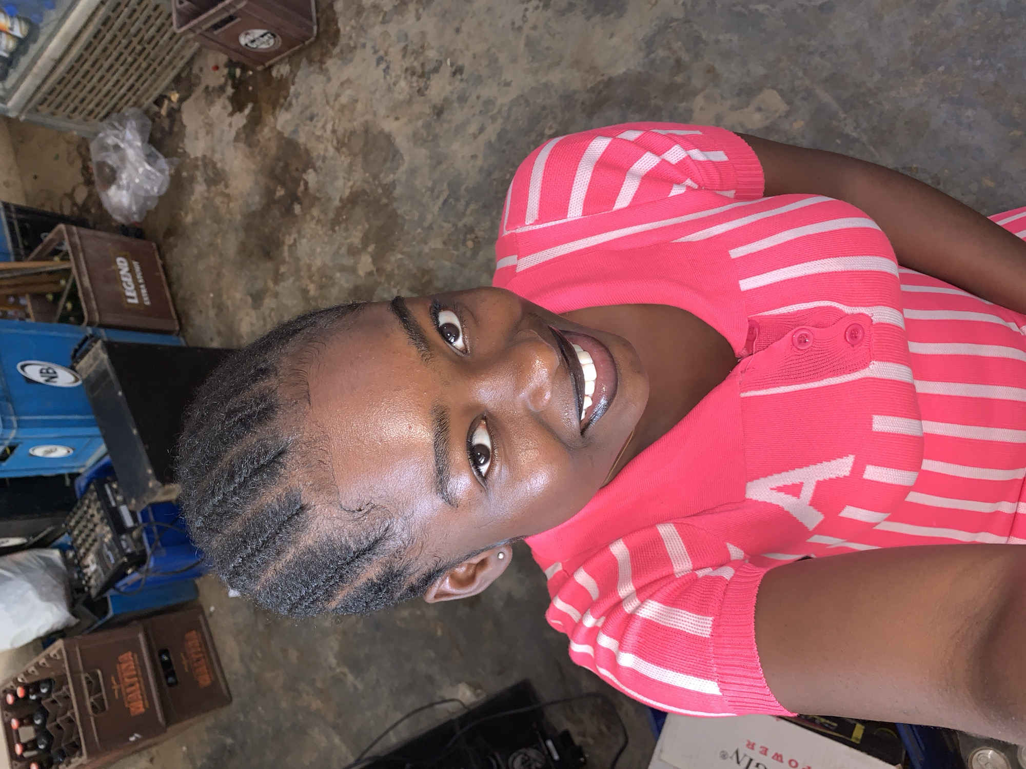 The image shows a woman with braided hair, wearing a pink and white striped top with buttons, looking directly at the camera and smiling. She appears to be indoors, surrounded by various objects including beverage crates labeled "LEGEND" and "MALTINA," and other unidentified equipment or containers. The flooring appears to be concrete. The location is suggested to be Kabong, Nigeria, based on external context. There are no specific actions or interactions visible beyond the woman's direct gaze at the camera. The lighting conditions are somewhat dim.