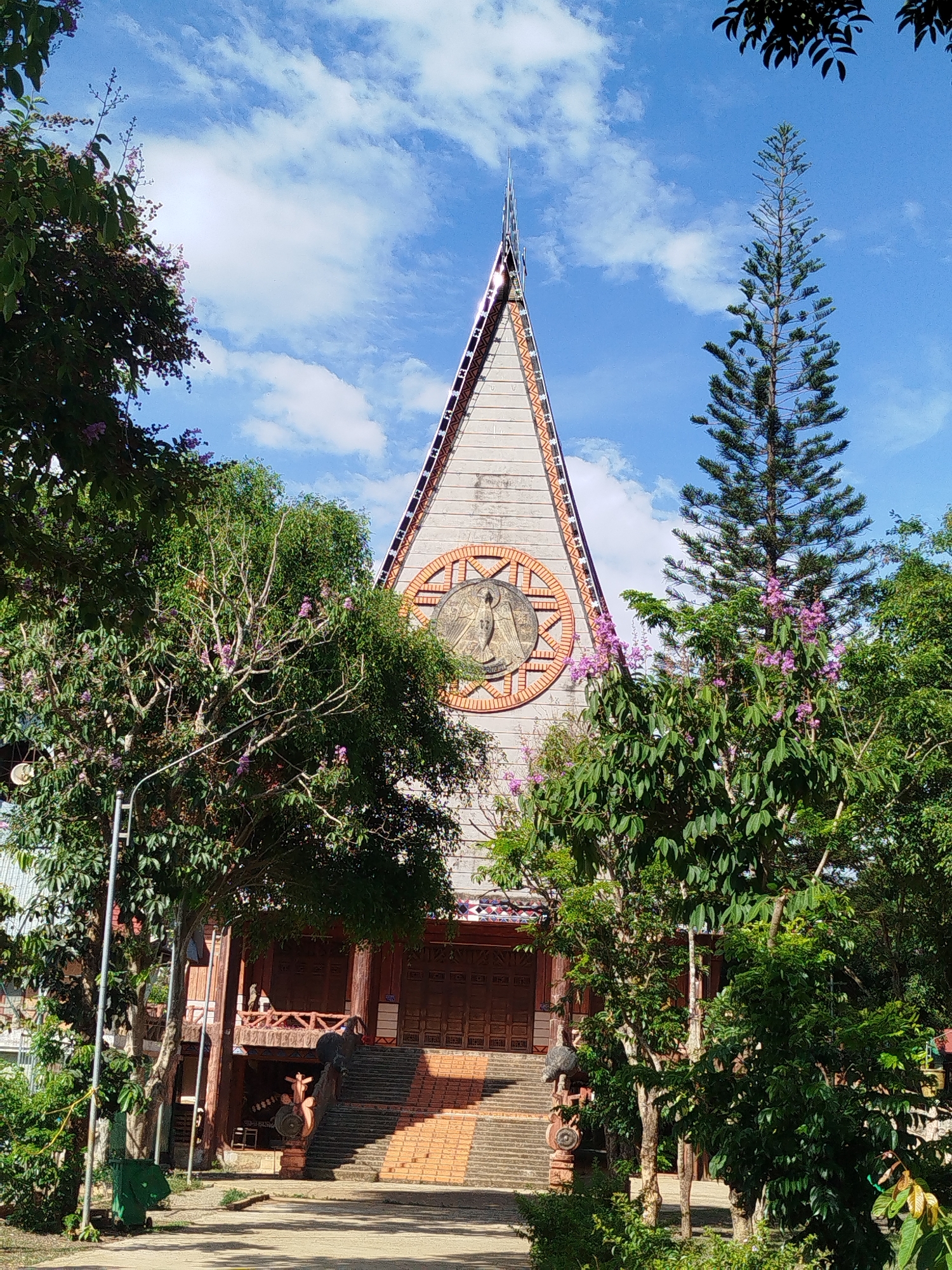The image depicts a building with a unique architectural design in An Phu Ward, Vietnam. The building has a tall, triangular facade with a central circular motif featuring a religious figure. A series of steps lead up to the building's entrance. Trees and foliage partially obscure the view of the structure. The sky above is blue with scattered clouds. A green trash can is visible on the left side of the image.