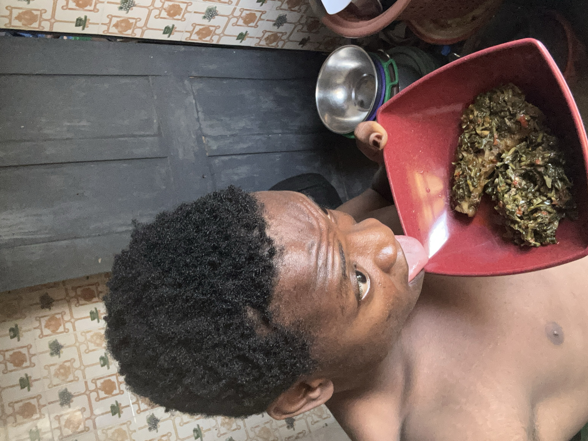 A medium shot depicts a Black man with dark hair eating from a red plate. He is shirtless. The plate contains a serving of leafy greens. In the background, there's a wooden door, a silver bowl, and a tiled floor. The location is Unknown, Unknown.