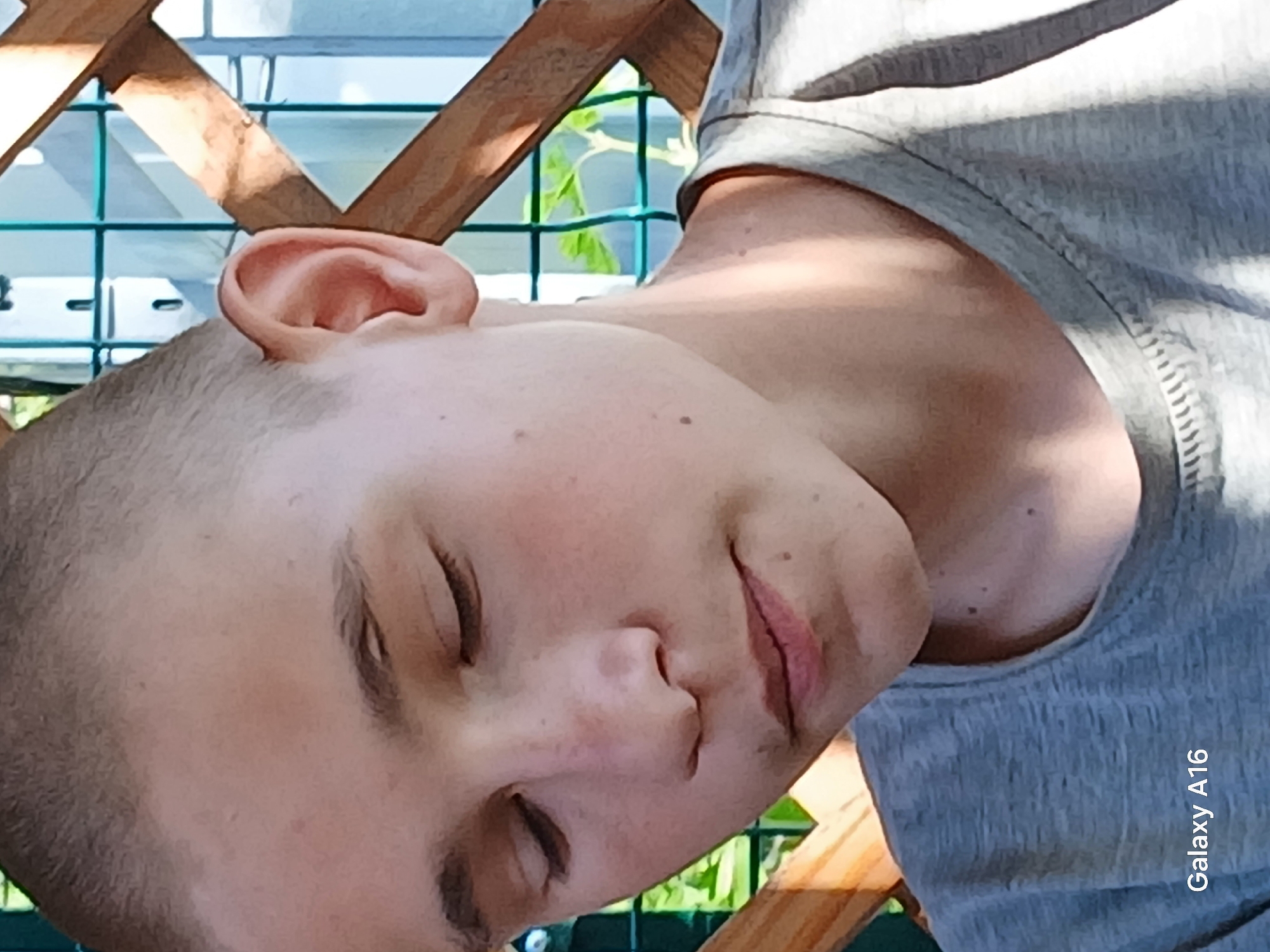This is a close-up, sideways, over-the-shoulder shot of a young person with a shaved head, resting outdoors. The subject's eyes are closed, and their head is tilted, suggesting they might be sleeping or relaxing. The lighting indicates it is daytime, with sunlight casting shadows. The background features a wooden structure, possibly part of a fence or a chair, and a green mesh fence with foliage visible behind it, suggesting a garden or outdoor recreational area. The person is wearing a gray t-shirt. The visible text on the bottom right of the image reads "Galaxy A16", likely indicating the device used to capture the photo. The overall mood is peaceful and serene.
