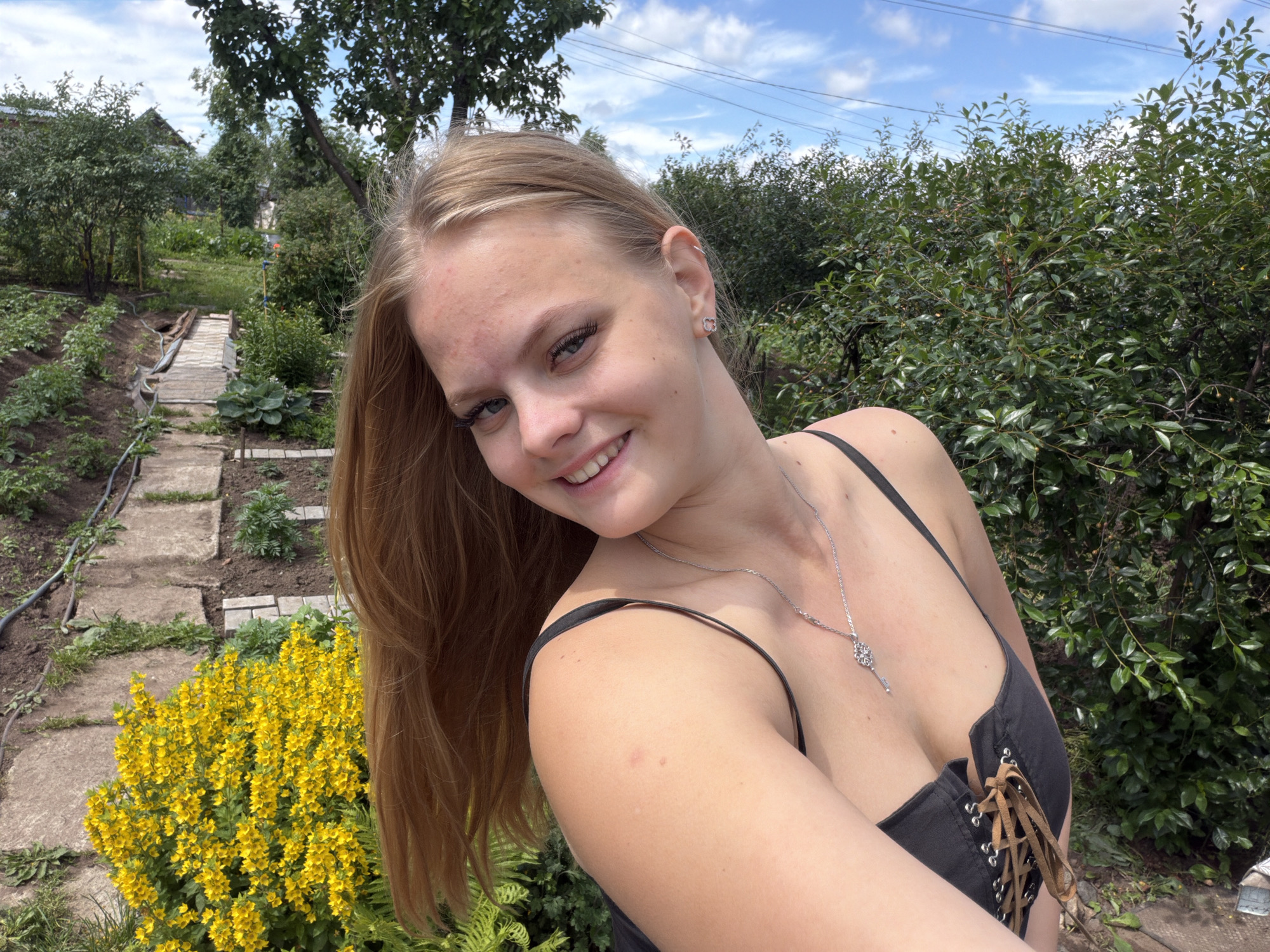 In this medium shot, a young woman with long blonde hair and light skin takes a selfie outdoors. She is smiling, with her head tilted slightly to her left. She is wearing a black tank top with spaghetti straps and a black corset-style top with brown lacing. A silver necklace with a key-shaped pendant rests on her décolletage. She has on heart-shaped earrings.
The background shows a garden or allotment with rows of plants and a paved walkway. Bright yellow flowers are blooming prominently in the foreground to the left of the woman. Lush green bushes and trees are visible behind her, and the sky above is blue with scattered white clouds. The overall lighting suggests it is daytime, likely a sunny day. The presence of cultivated plants and trees implies a rural or suburban setting, and the crops provided confirm the location as Chaikovsky, Russia. The woman's expression and the bright, natural setting evoke a relaxed and happy mood. There is no visible text in the image.