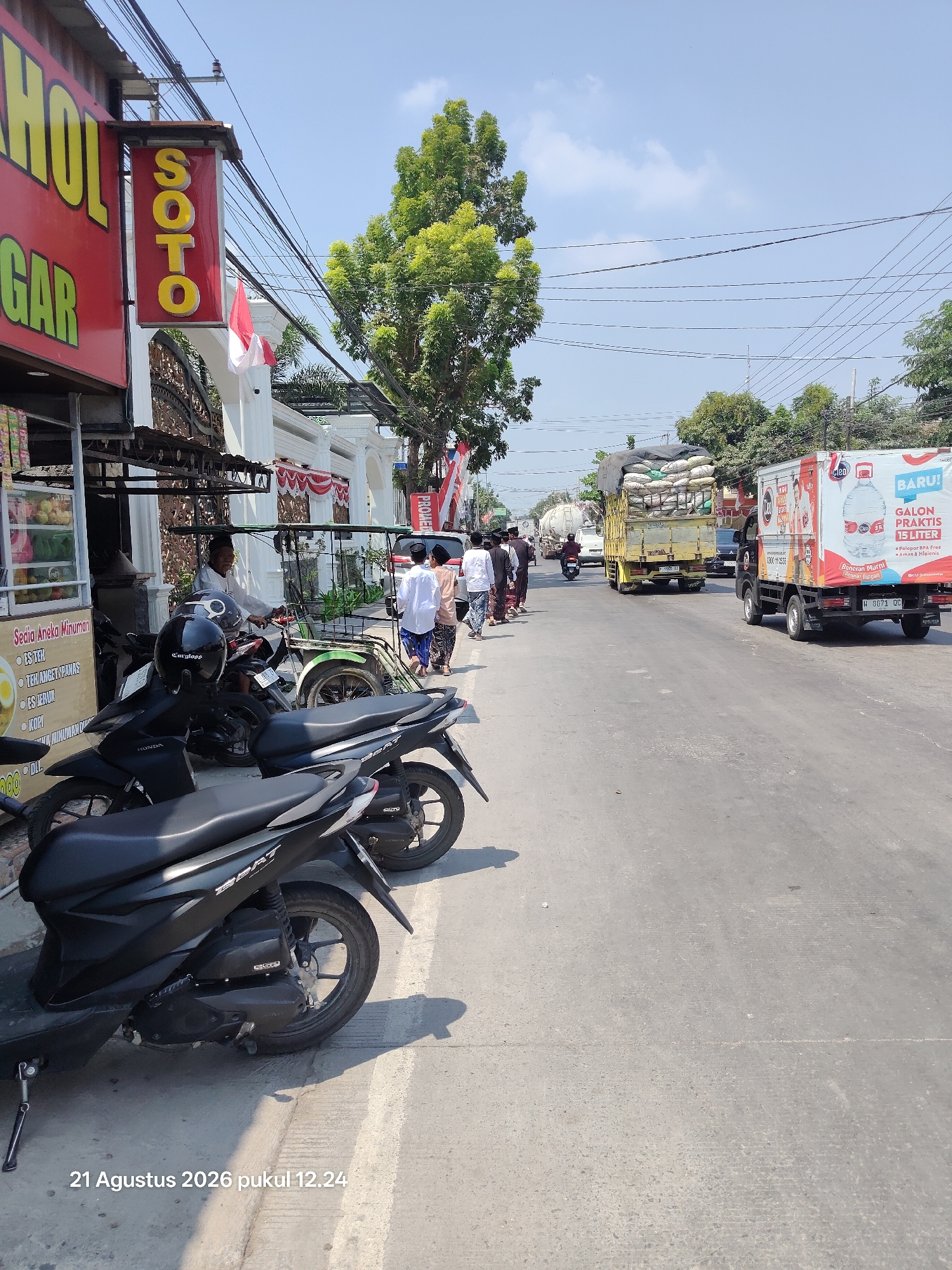 The image captures a street scene in Kalitidu, Indonesia on August 21, 2026, at 12:24 PM. In the foreground, several parked motorcycles line the side of the road. A food stall with a red awning is visible on the left, displaying various items and signs in Indonesian. A prominent red sign with yellow lettering reads "SOTO," indicating a local eatery.
Further down the street, a group of people, some wearing traditional Islamic attire, are walking. A man is seen riding a pedal-powered vehicle. Traffic is present, including a yellow truck loaded with sacks and a white delivery truck with prominent branding. Electrical wires are strung overhead, typical of a developing urban or semi-urban environment. The sky is partly cloudy. The overall atmosphere suggests a busy street in a commercial area.
